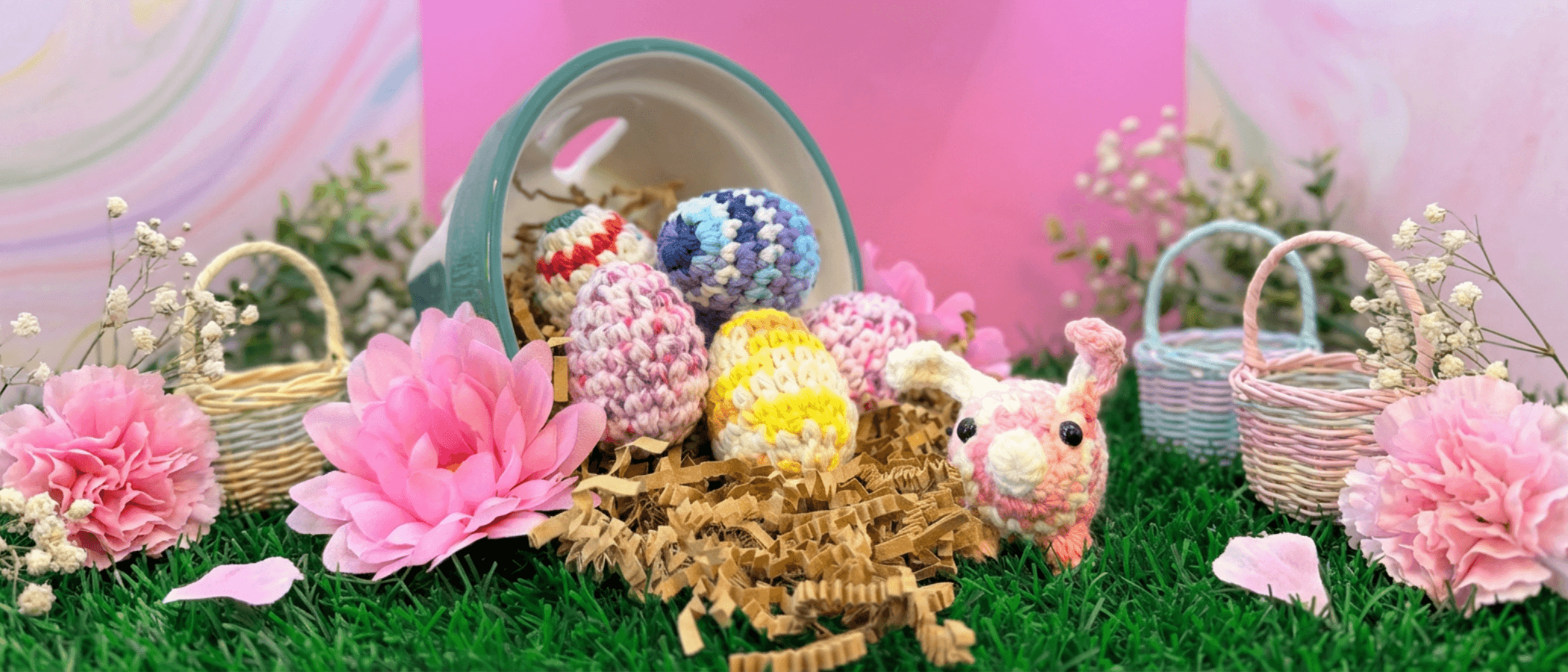 Pink crocheted bunny with white face sits beside six colorful hand-crocheted eggs in a teal basket filled with straw, surrounded by pink flowers and small decorative baskets on artificial grass.