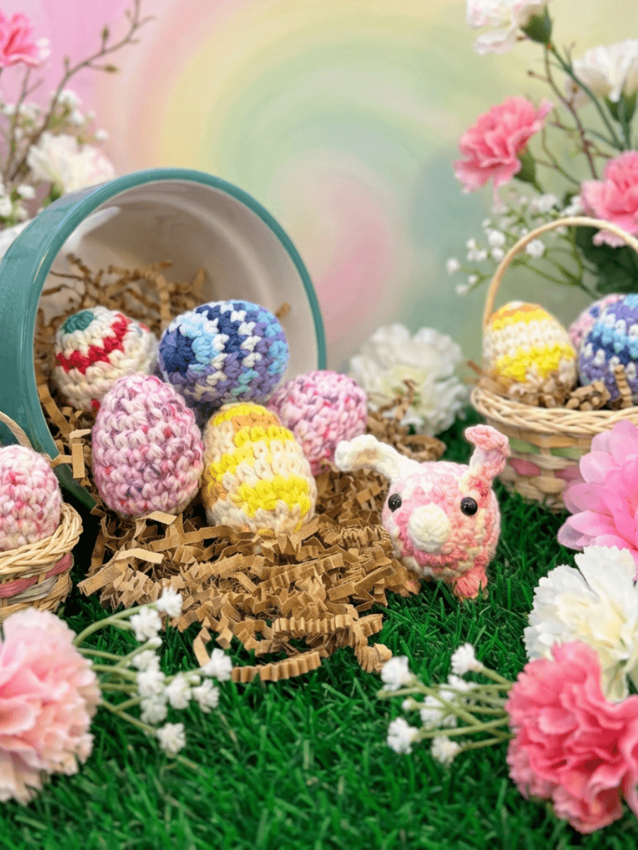 Crocheted pink and white striped bunny with black bead eyes sits on artificial grass beside colorful hand-crocheted eggs in pastel stripes, arranged with wicker baskets and pink carnation flowers.