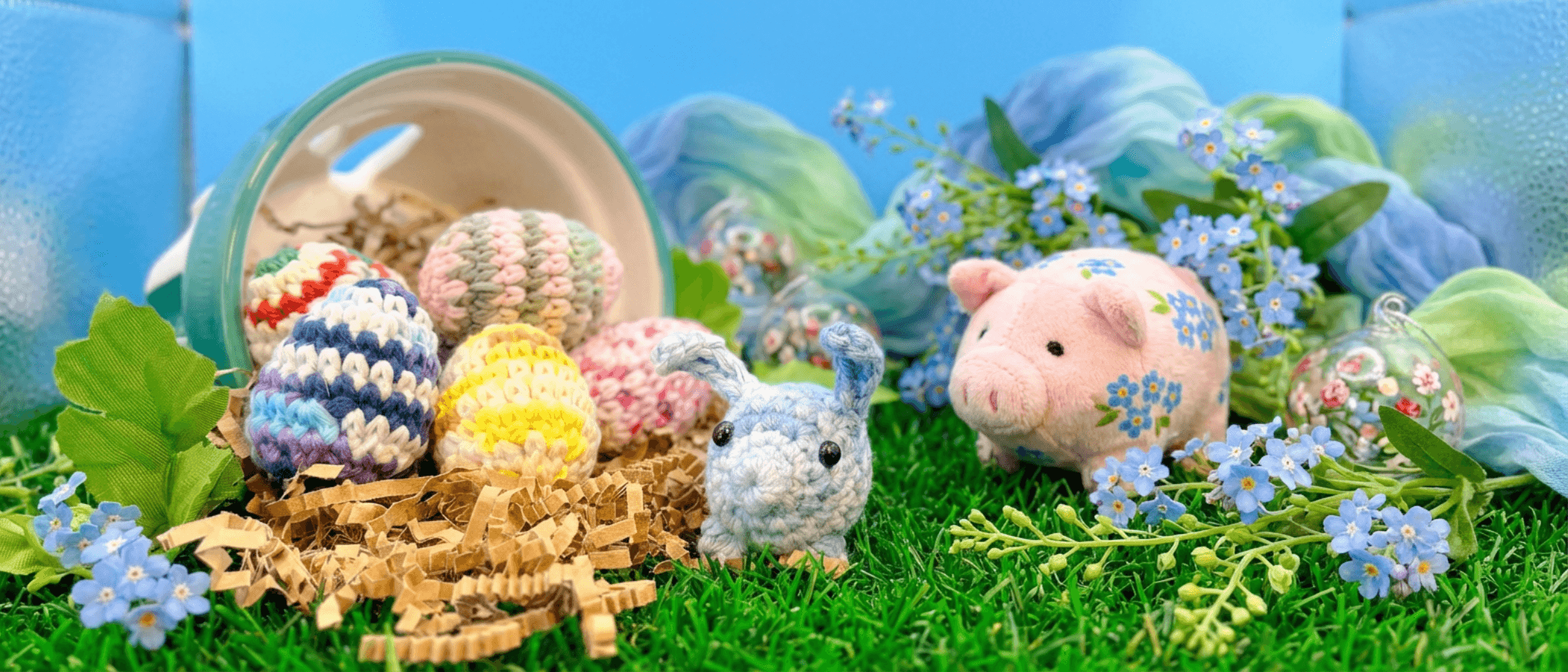 Pastel blue crocheted bunny with black bead eyes sits on artificial grass beside six striped crocheted eggs, pink plush pig, and blue forget-me-not flowers against blue backdrop.