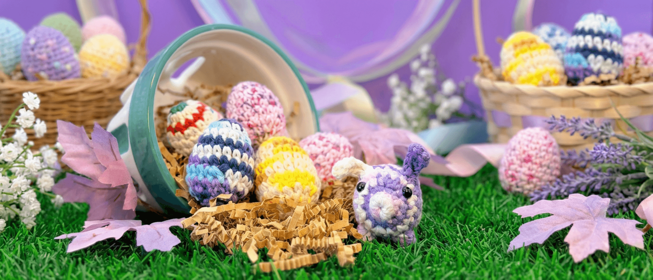 Easter scene with colorful hand-crocheted eggs spilling from a tipped basket, a purple bunny plush, dried grass, silk flowers, and lavender stems arranged on artificial green grass.