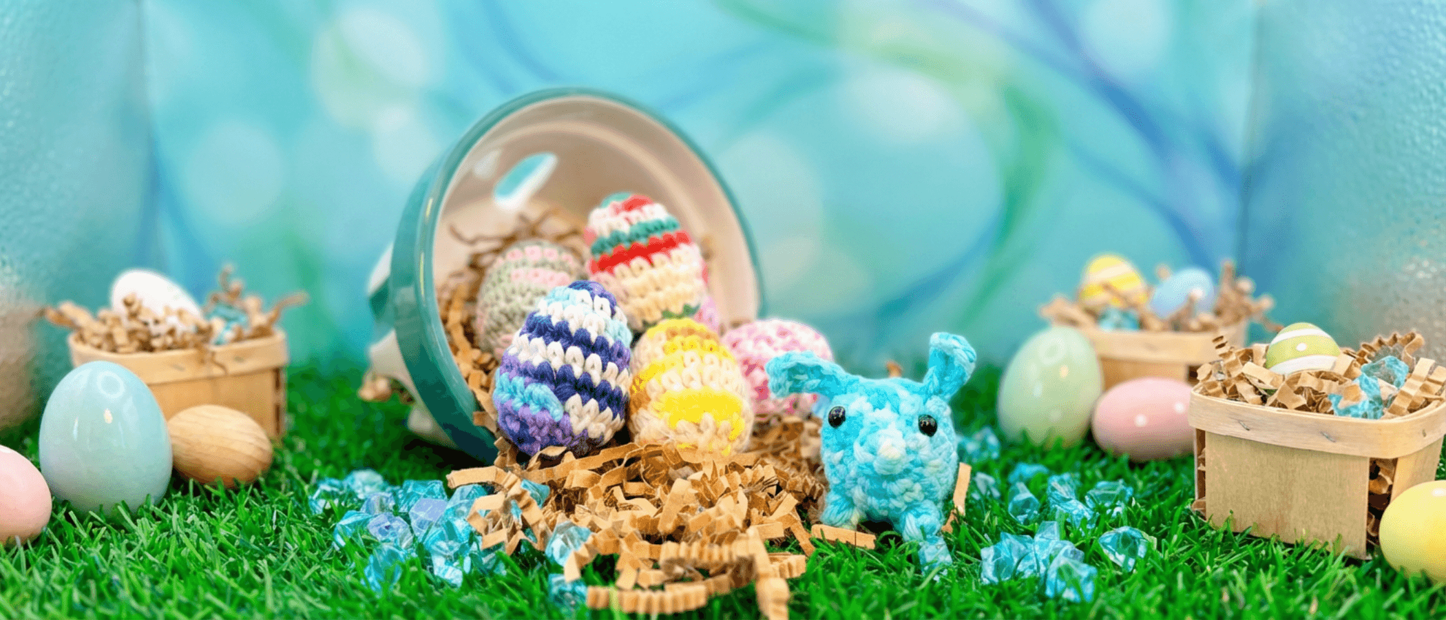 Tipped-over teal basket with colorful hand-crocheted Easter eggs spilling onto artificial grass alongside a small turquoise crocheted pig character and wooden crates filled with shredded paper.