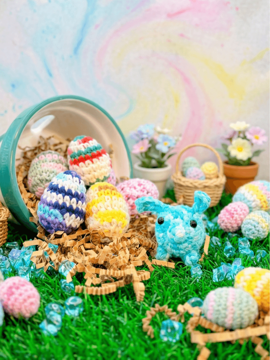 Crocheted turquoise bunny and colorful striped Easter eggs spilling from teal bucket onto artificial grass. Handmade amigurumi display with basket and flower pots in background.