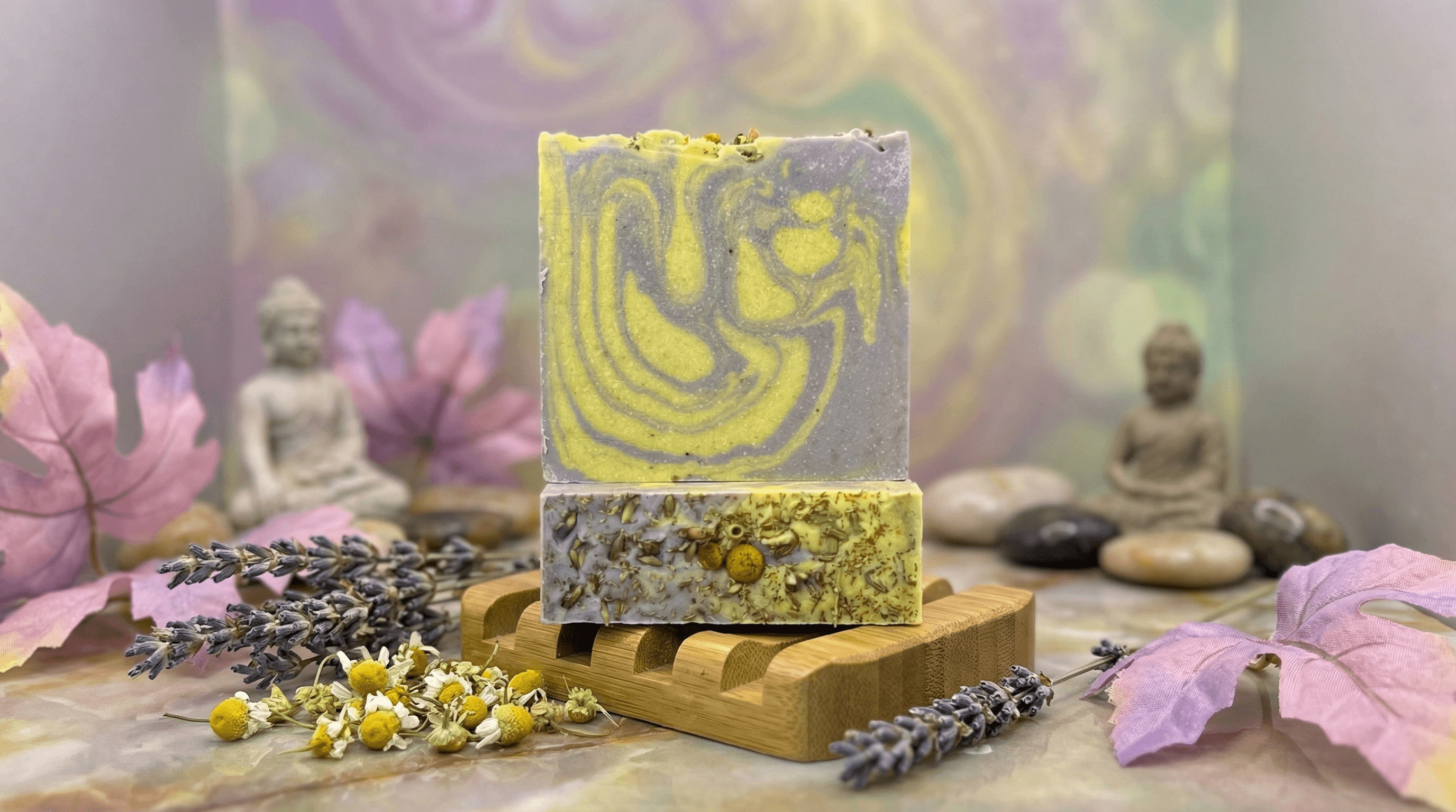 Two stacked handmade soap bars on bamboo dish with swirled yellow and gray design on top, chamomile flowers and lavender sprigs visible in bar and surrounding display. Buddha statues and lotus flowers blurred in background.