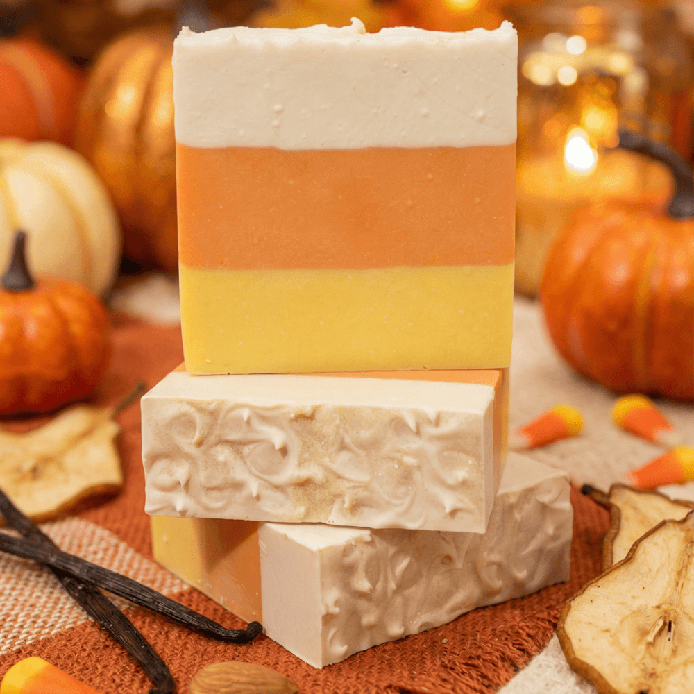 Stack of handcrafted soap bars in candy corn colors: yellow, orange, and white horizontal layers. Top bar shows smooth surface, bottom bars feature decorative swirl patterns on top.