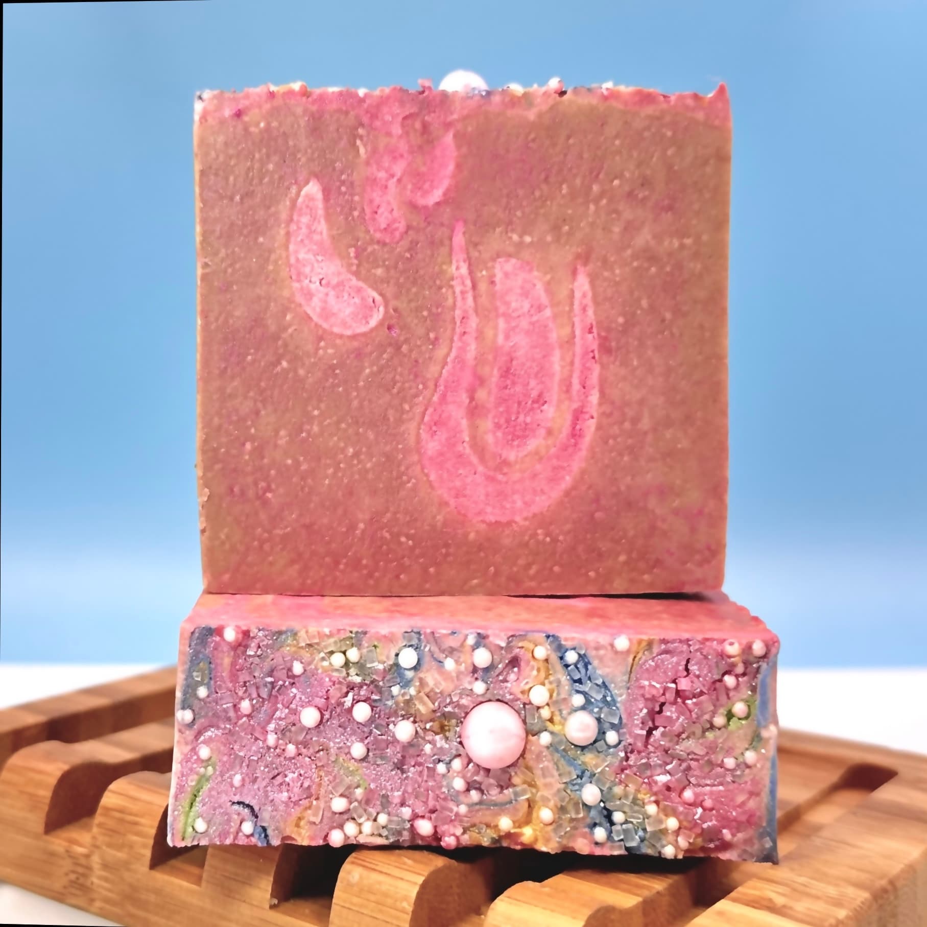 Two pink and brown marbled soap bars stacked on a wooden dish. The top bar has a swirled design, while the bottom bar is embedded with white, pink, and gold spherical beads.