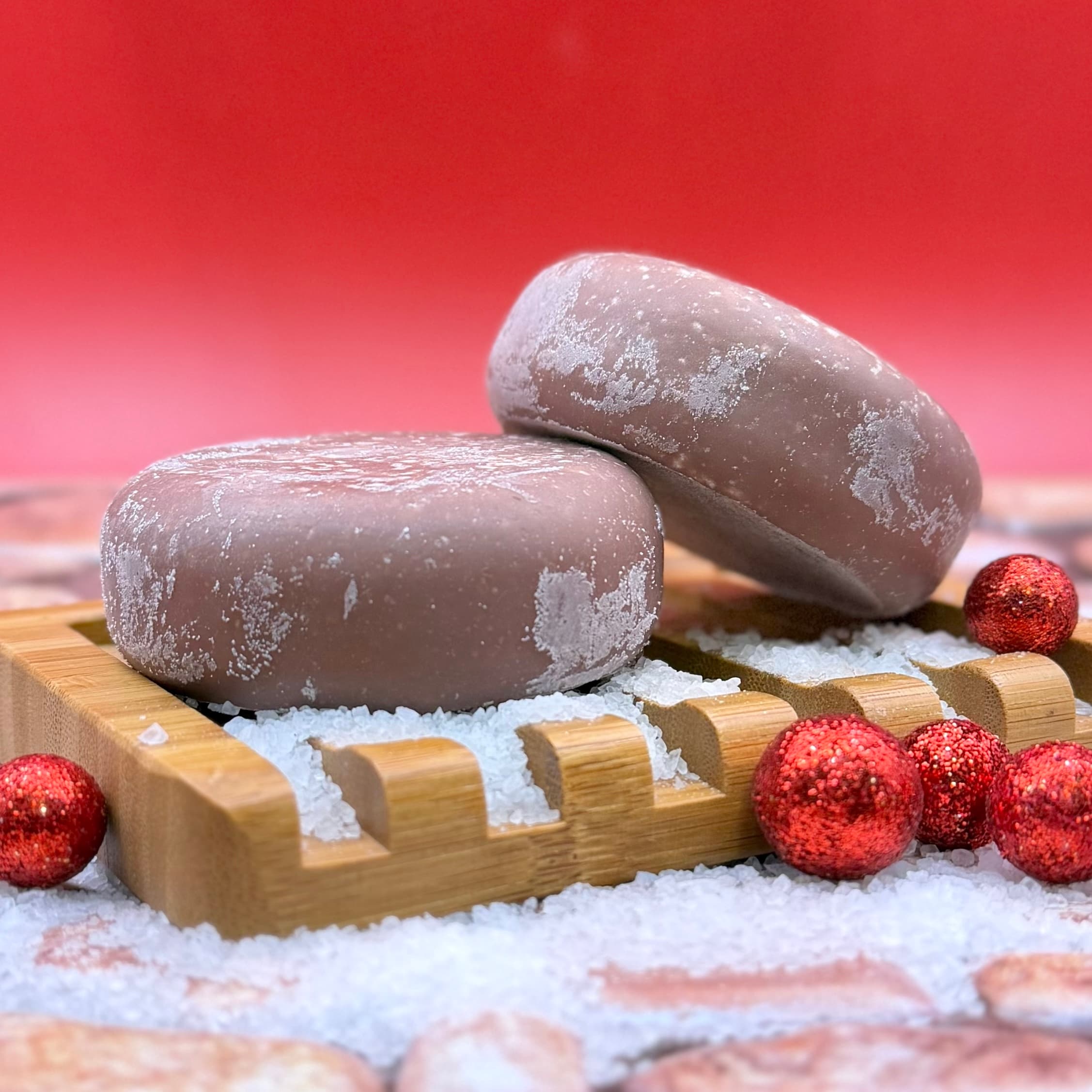 Two round mauve-gray bars dusted with white coarse sea salt, stacked on a wooden soap dish surrounded by snow and red glitter ornaments against a pink gradient background.