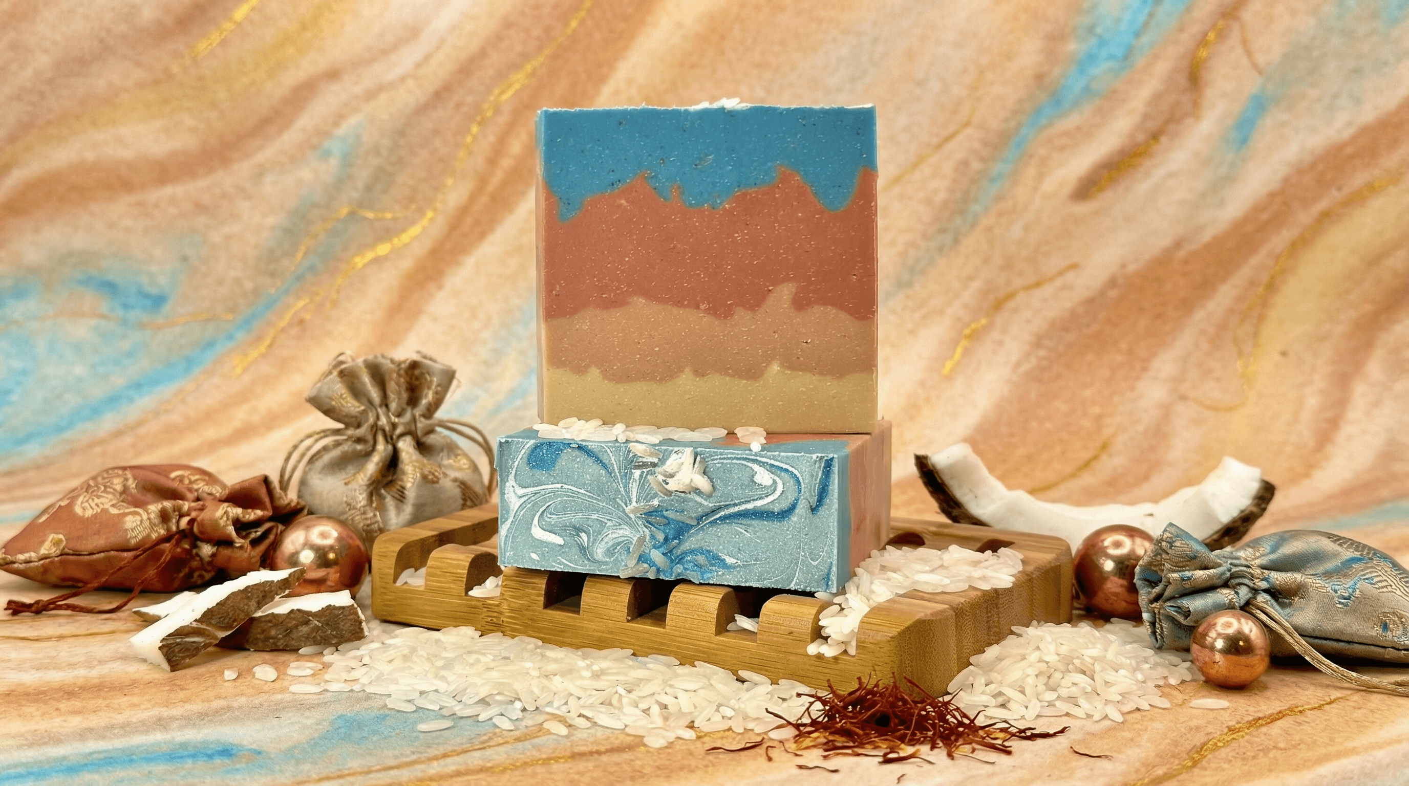 Two handcrafted soap bars on wooden pallets: one with marbled blue and sage swirls, another with desert sunset layers in blue, rust, sand, and cream. Styled with rice grains, saffron, silk pouches, and ornaments on a swirled backdrop.