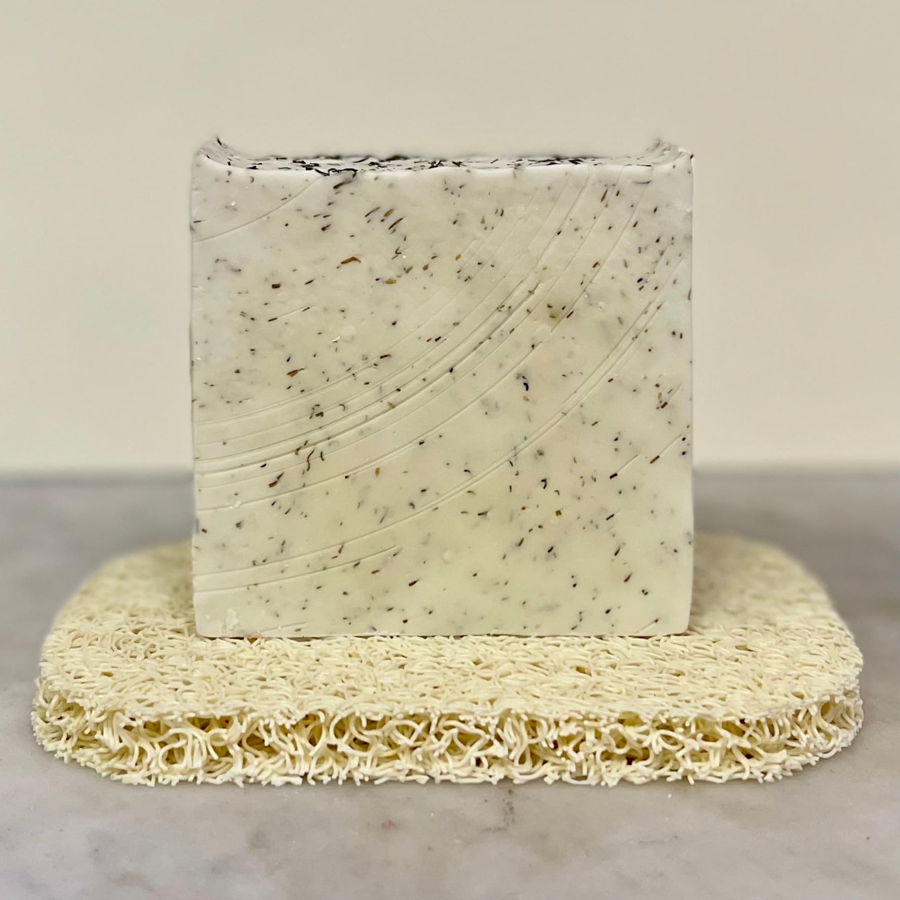 Cream-colored handmade soap bar with dark speckles and diagonal swirl pattern, displayed on a beige textured soap saver pad against a neutral background.