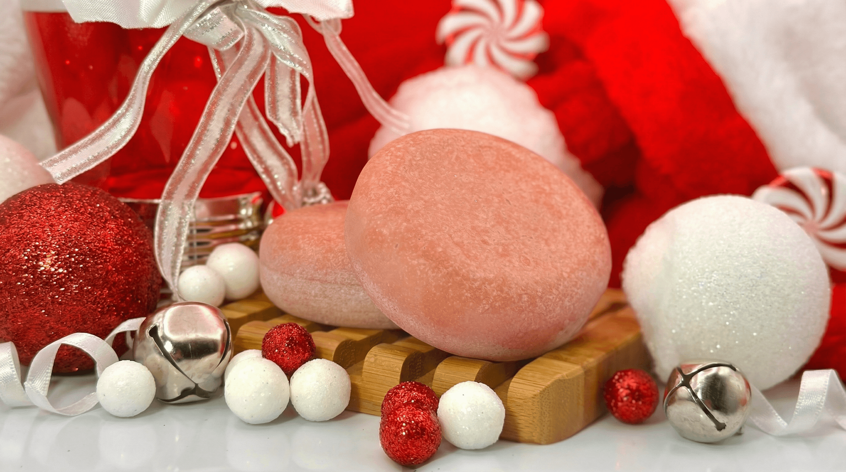 Pink heart-shaped sea salt scrub bar with sparkly sugared texture on wooden soap dish, surrounded by red and white holiday ornaments, silver bells, and peppermint-striped decorations.