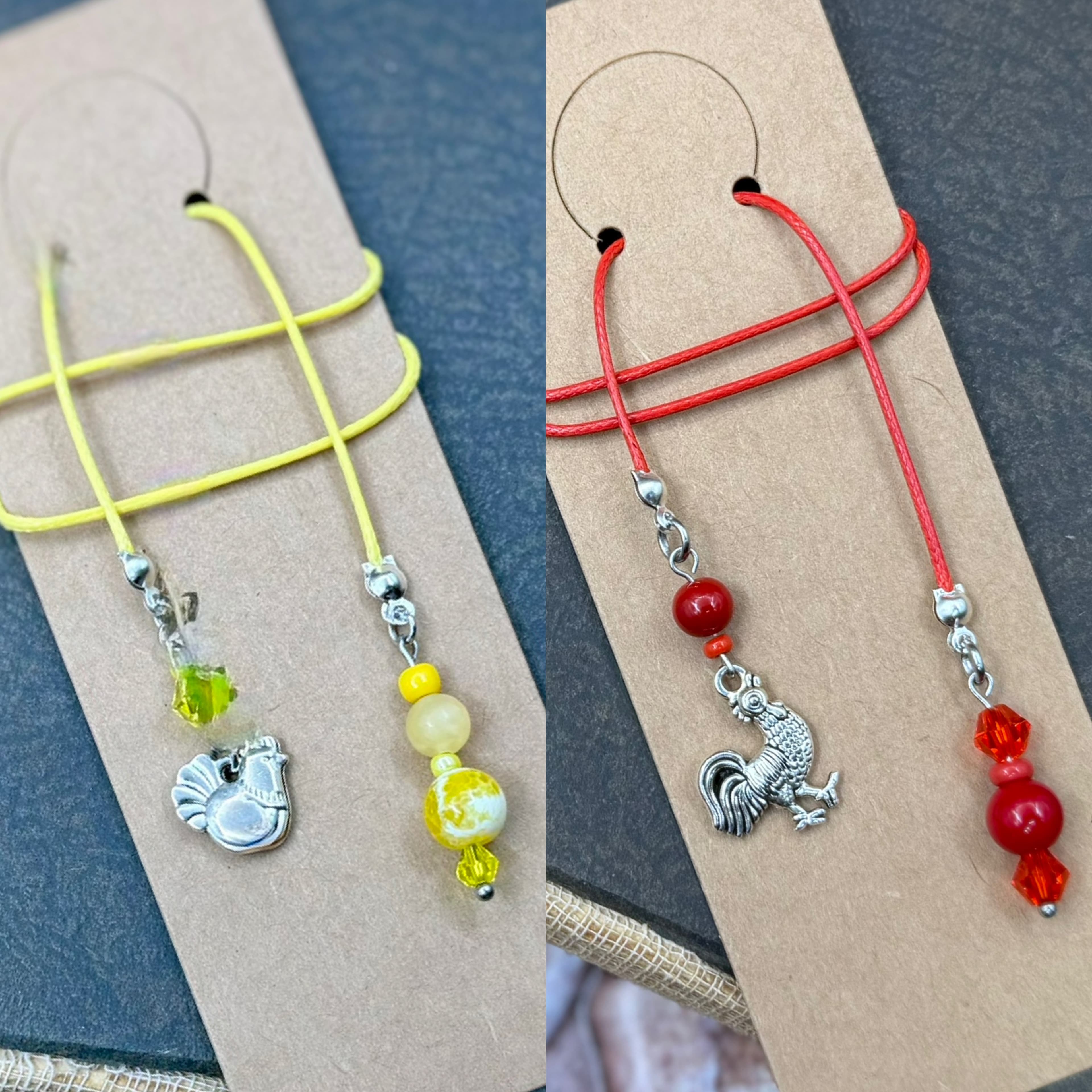 Two handmade bookmarks with yellow and red cords featuring rooster and hen charms with coordinating beads, displayed on kraft paper cards against a blue textured background.