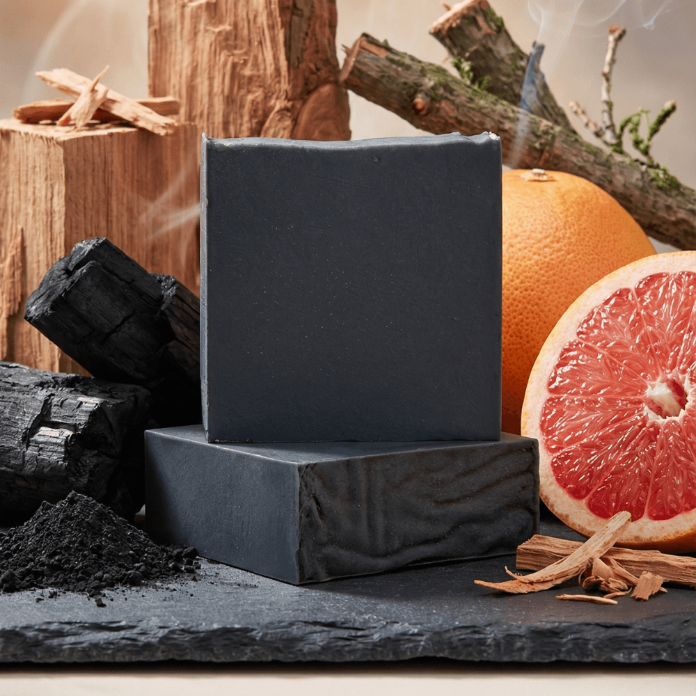 Dark charcoal-gray soap bars with smooth matte finish, displayed with chunks of black activated charcoal, fresh grapefruit halves, and rustic wood pieces on slate surface.