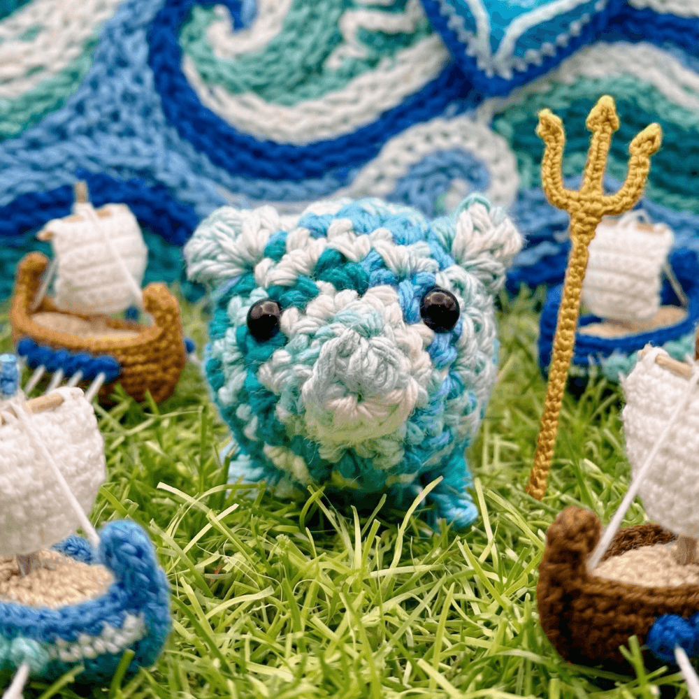 Hand-crocheted blue and white variegated pig with black bead eyes and golden trident, positioned on artificial grass with ocean wave blanket and small sailboat props in background.
