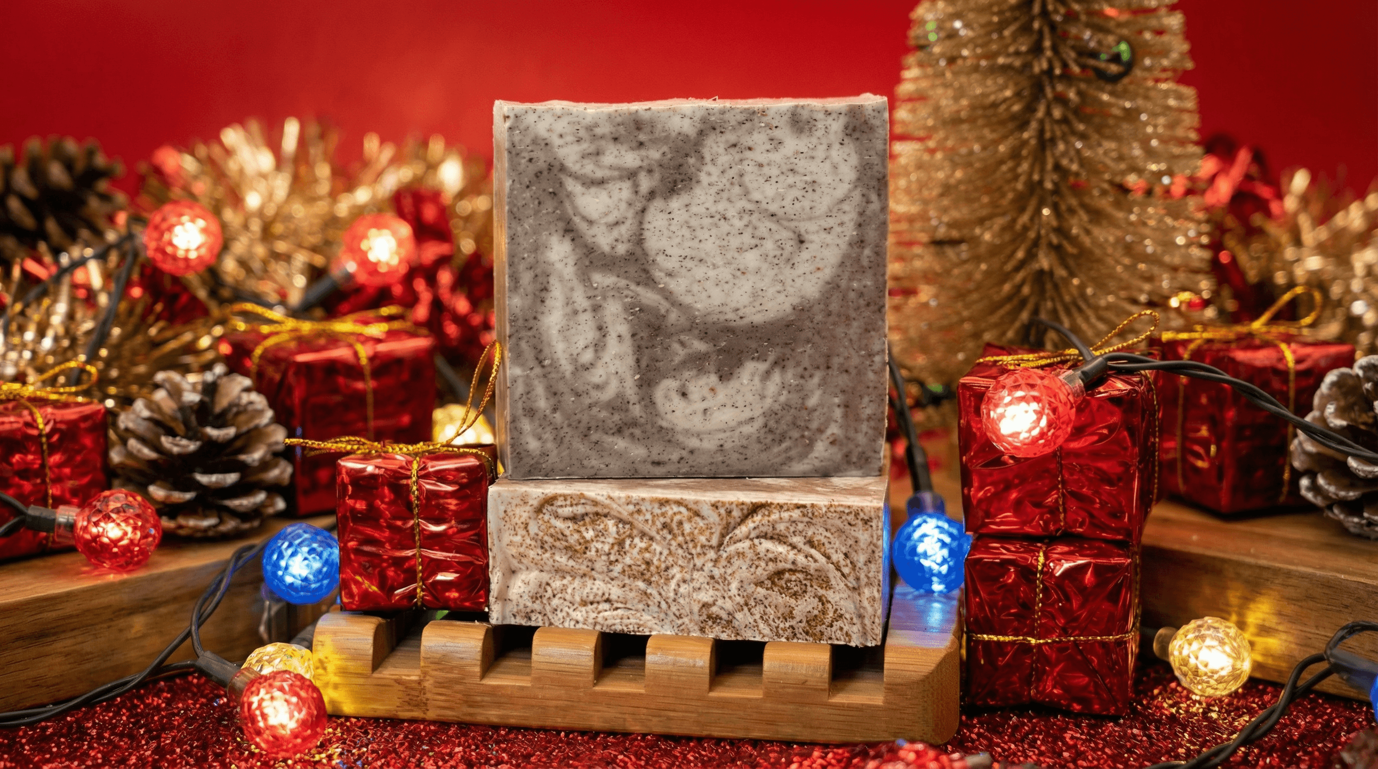 Two bars of speckled grey and brown swirled soap stacked on a wooden soap dish, surrounded by festive holiday decorations including lit string lights, pine cones, and miniature gift boxes.