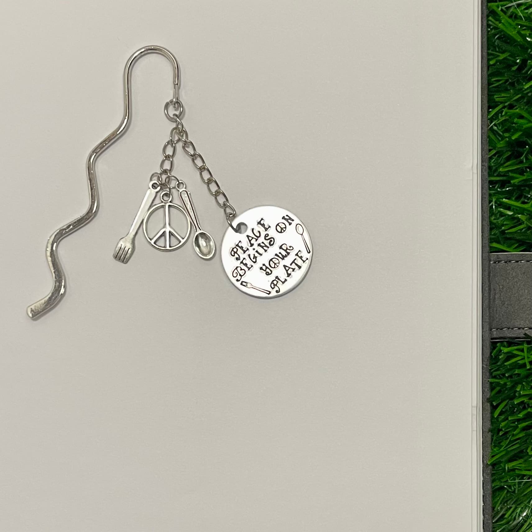 Silver bookmark with wavy design featuring a chain with hanging charms including a peace sign, fork, spoon, and stamped circular disc reading 'Peace Begins On Your Plate'.