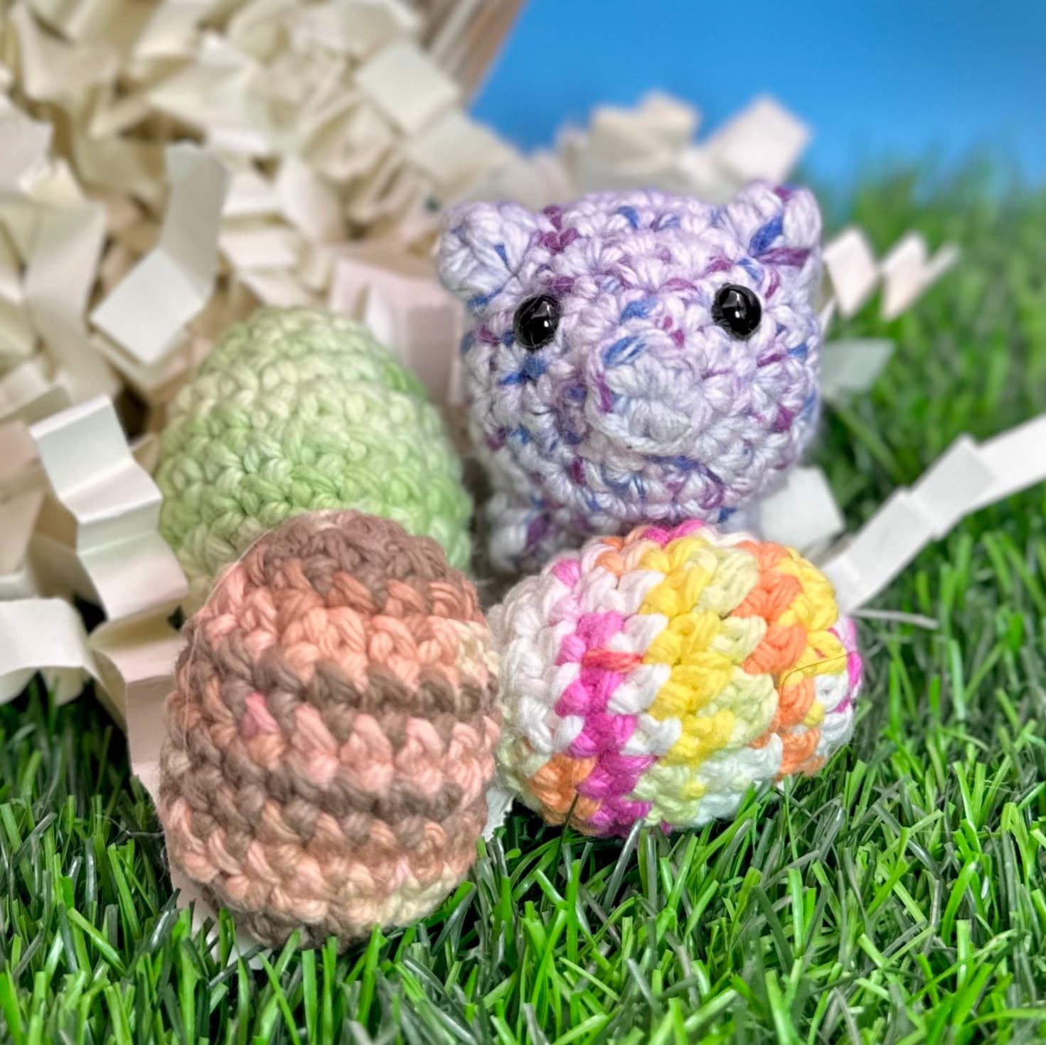 Four crocheted eggs in pastel colors (purple marbled, green, tan, and rainbow striped) rest on artificial grass with curled paper shredding. A small purple crocheted pig with black bead eyes sits behind them.