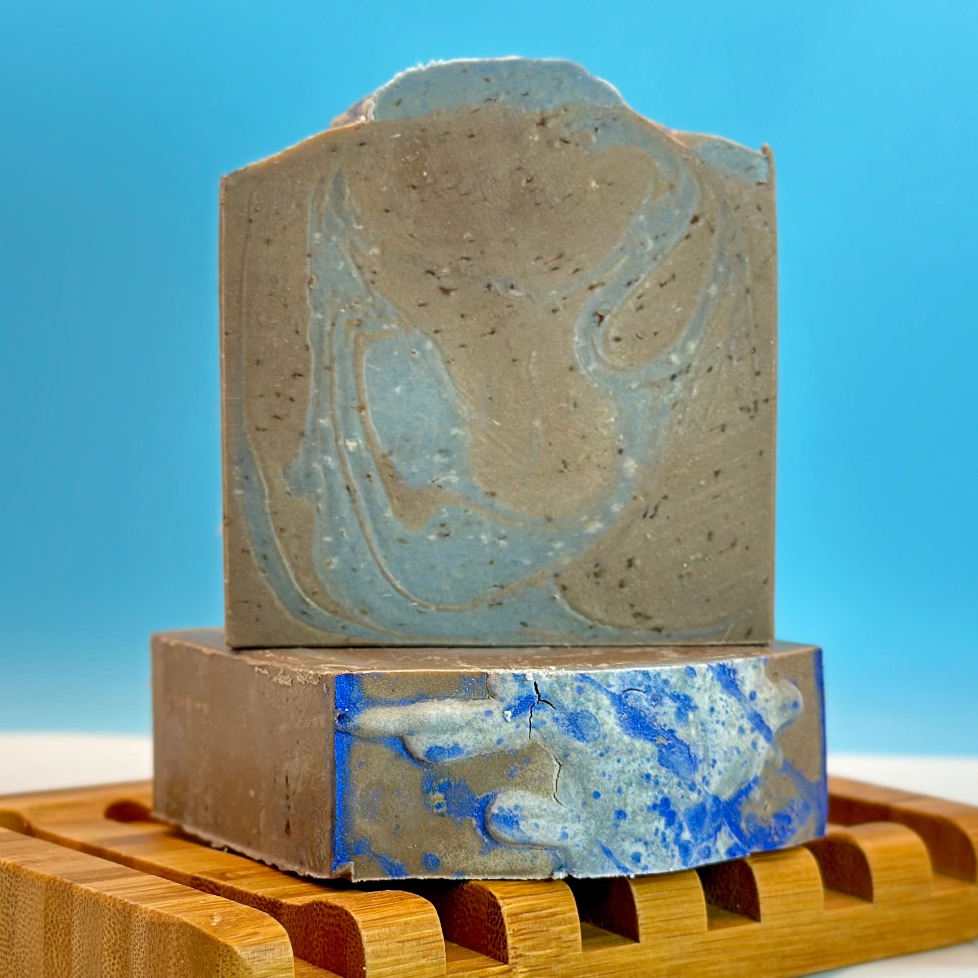 Two bars of beige soap with blue swirl designs stacked on a bamboo soap dish against a blue background. The top bar features an embossed leaf pattern while the bottom shows marbled blue streaks.