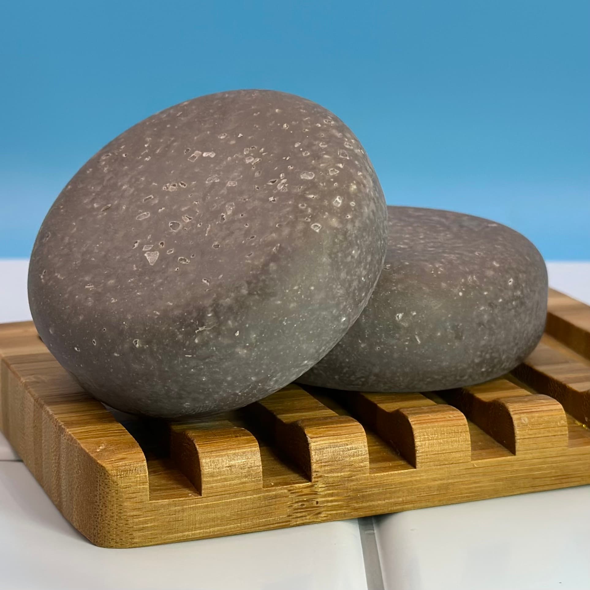 Two oval gray sea salt scrub bars with visible white salt crystals throughout, resting on a wooden soap dish against a light blue background.