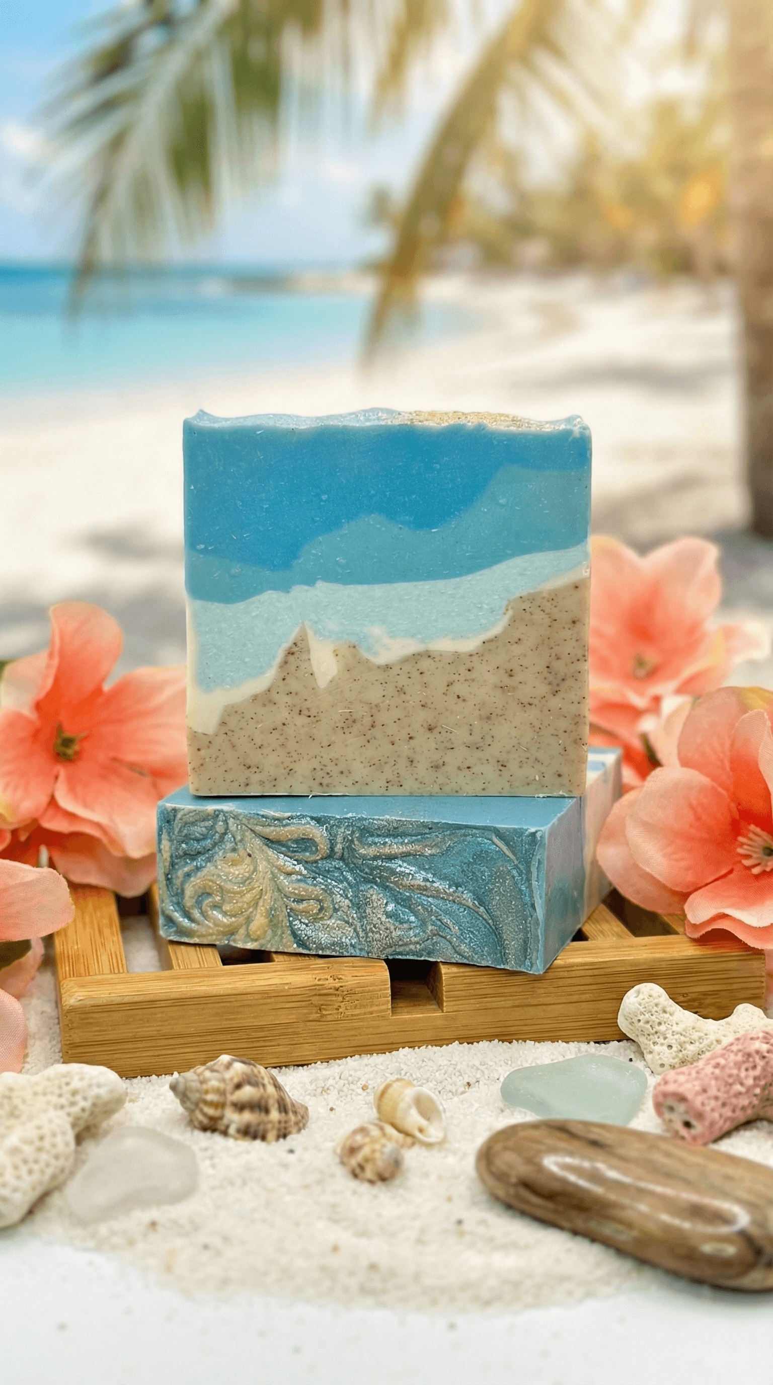 Two stacked handcrafted soap bars on bamboo tray with beach backdrop. Top bar shows layered ocean waves in blue tones over speckled sand-colored base. Bottom bar features swirled turquoise and gold pattern.