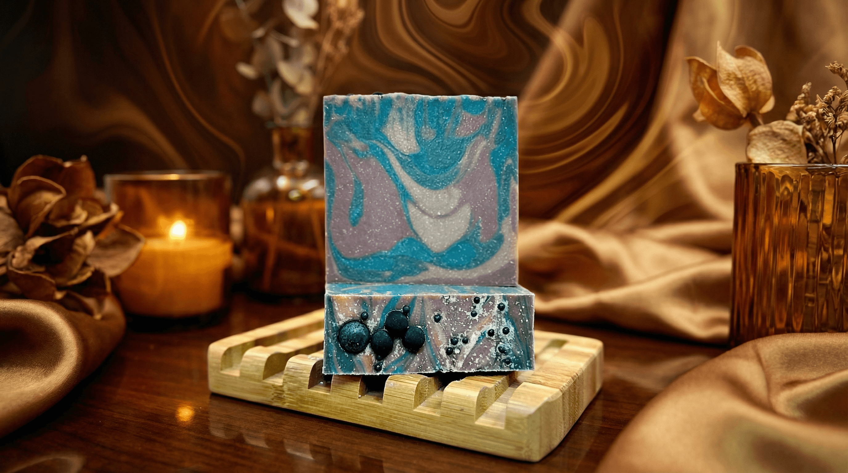 Two-toned handcrafted soap bar on wooden slat dish with swirled turquoise, lavender and white top, clear bottom half with embedded dark blue spheres. Warm candlelit setting with dried flowers.