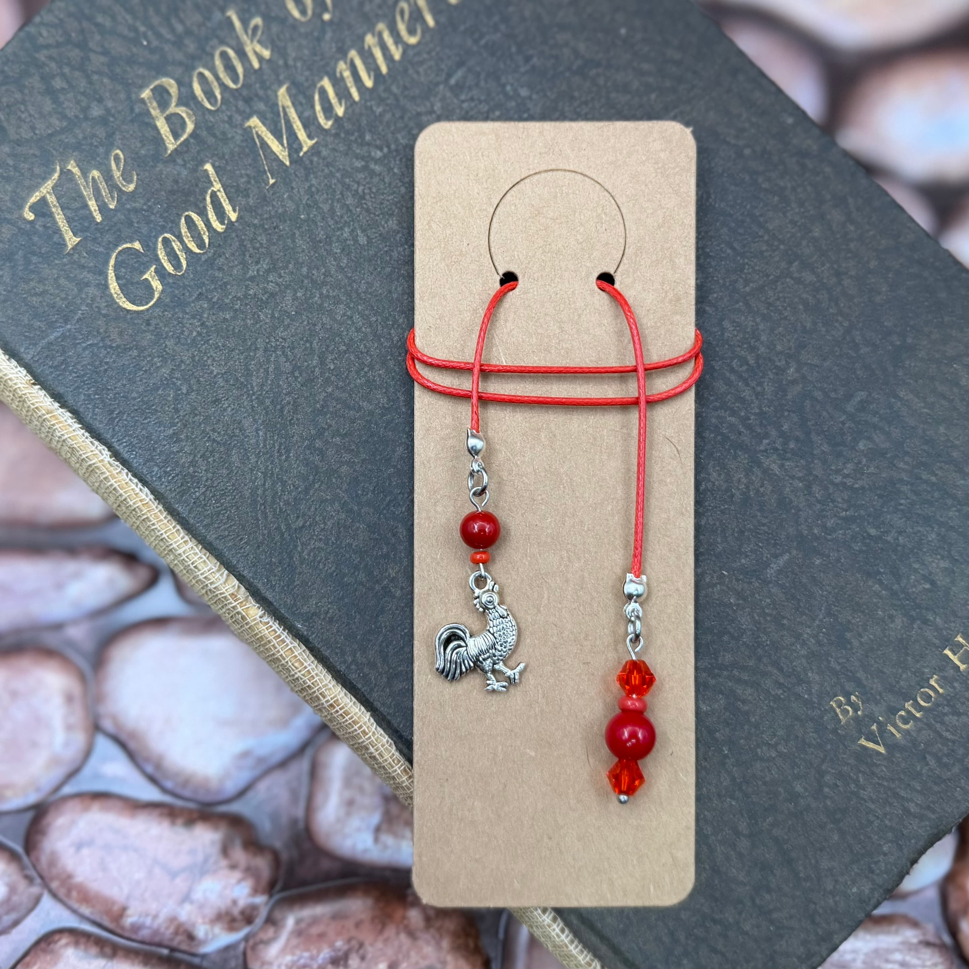 Handmade bookmark with red cord and rooster charm displayed on tan card, resting on 'The Book of Good Manners' with gold lettering. Red and orange beads accent the silver-toned charm.