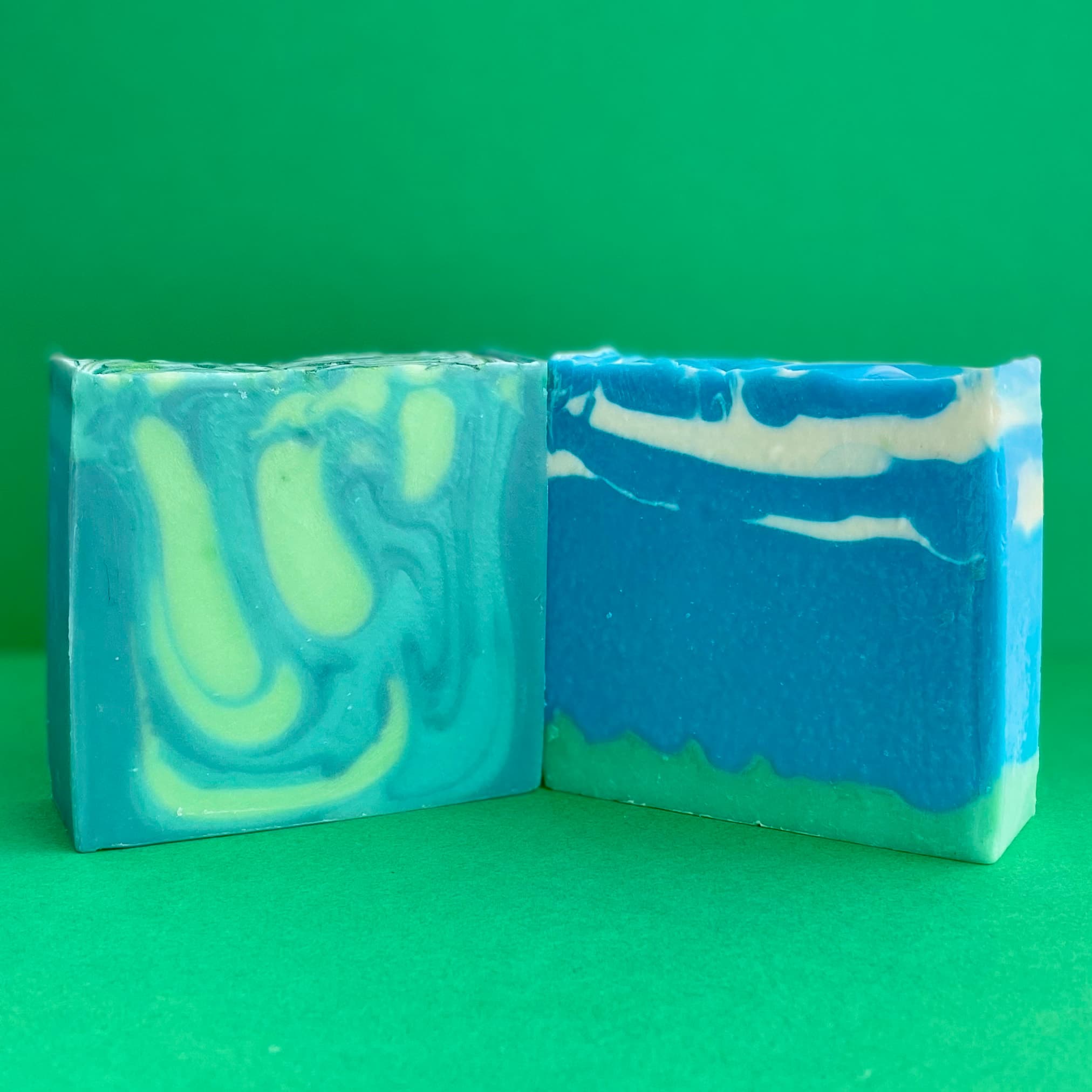 Two rectangular soap bars with swirled patterns in turquoise, bright blue, lime green, and white against a green background. The left bar features curved spiral swirls while the right has horizontal wave patterns.