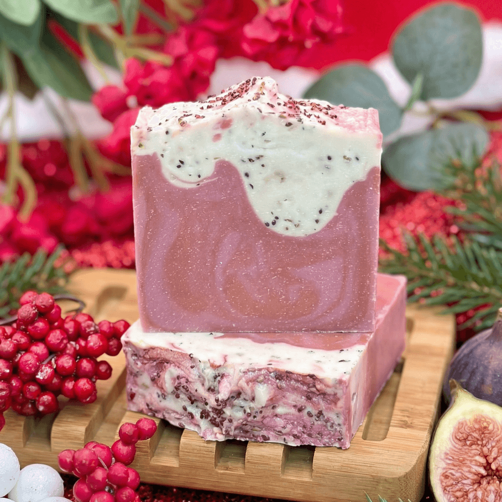 Two stacked handmade soap bars with pink-mauve base and white frosted tops decorated with dark speckles, displayed on bamboo tray with fresh cranberries, figs, and holiday greenery.