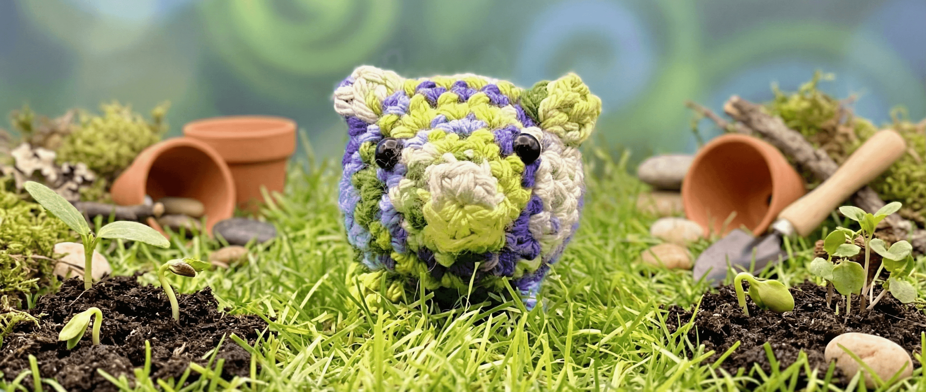 Hand-crocheted pig in yellow, purple, and green yarn sits on grass surrounded by terracotta pots, moss, seedlings, and small stones in a miniature garden scene.