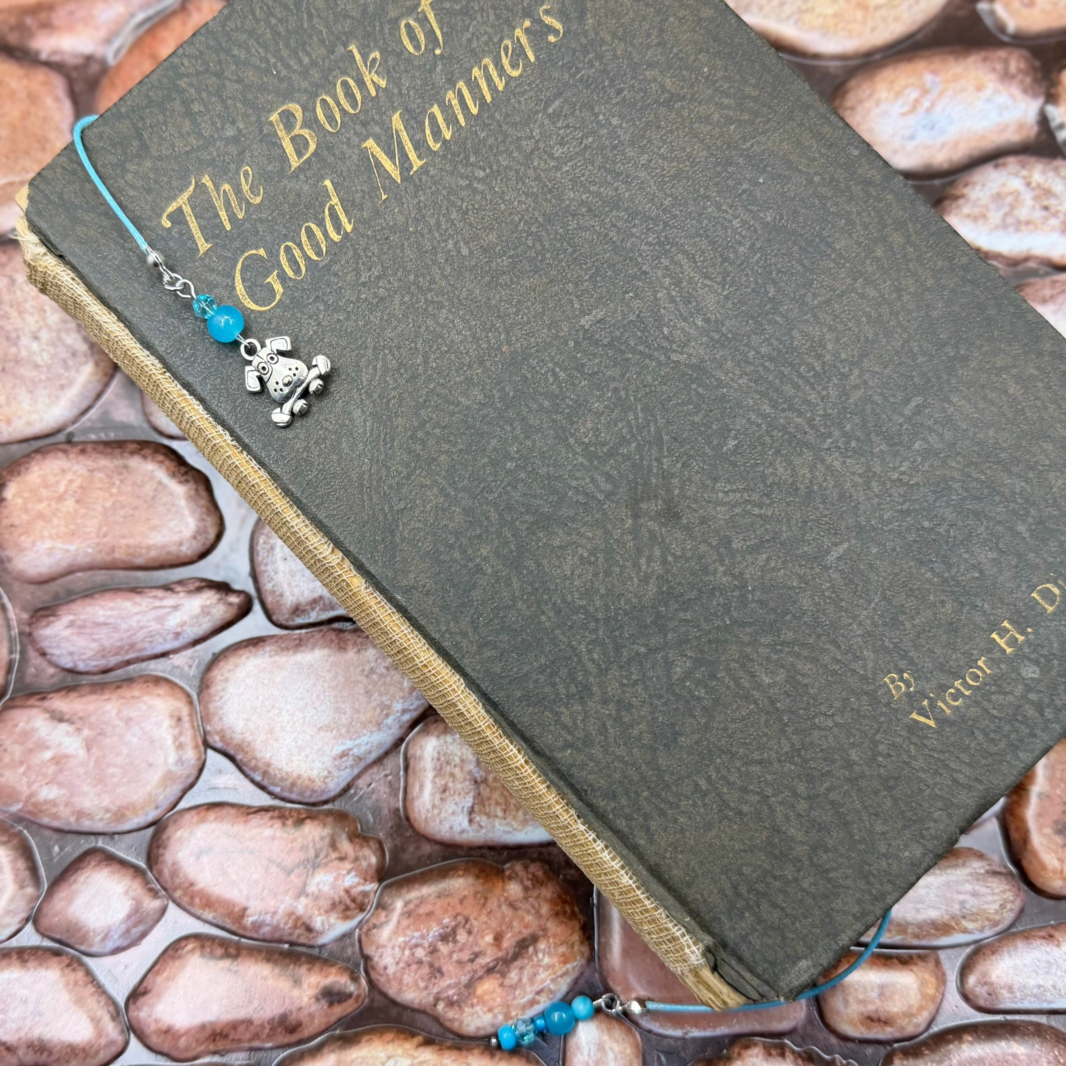 Gray hardcover book titled 'Book of The Good Manners' with gold lettering, adorned with a beaded bookmark featuring a silver dog charm and blue beads on tan cord, resting on smooth river stones.