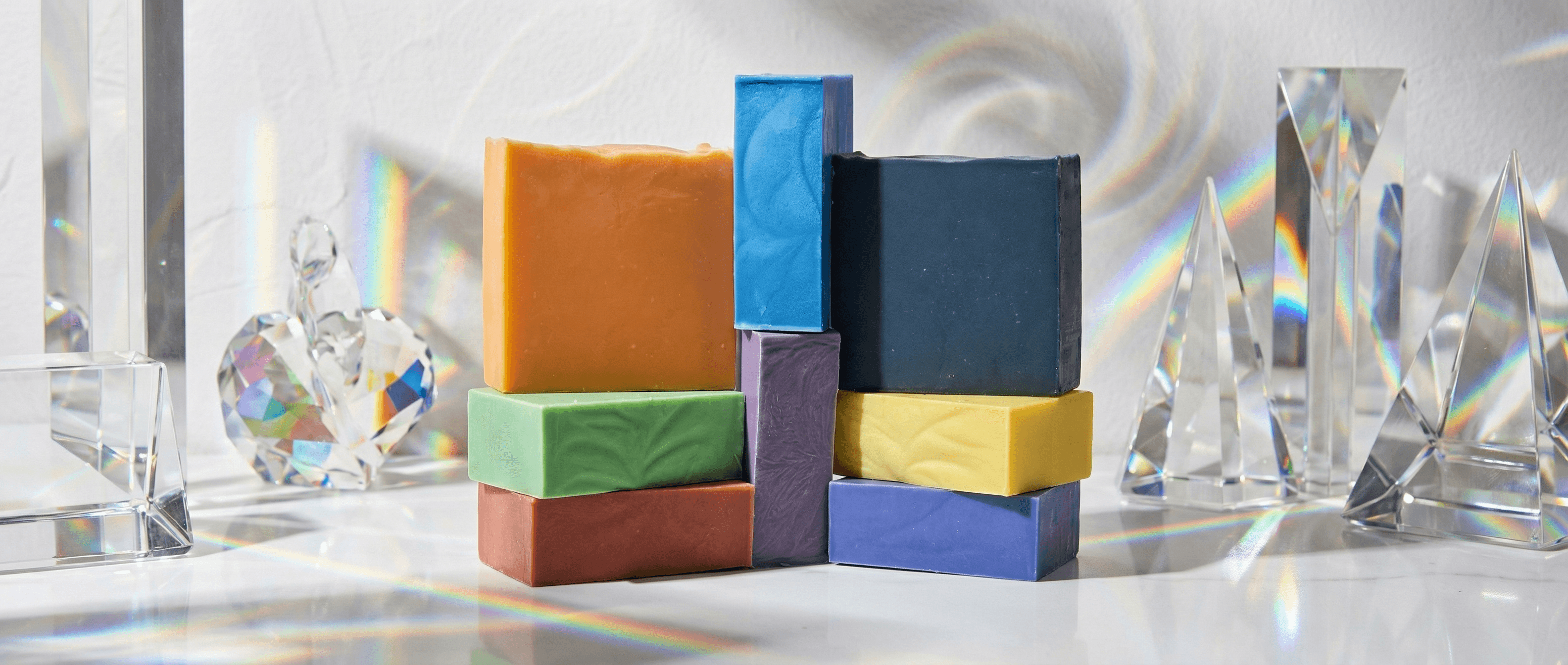 Eight rectangular soap bars in rainbow colors plus black, stacked in an architectural arrangement. Bars have matte, textured surfaces and are displayed among clear crystal prisms casting rainbow light.