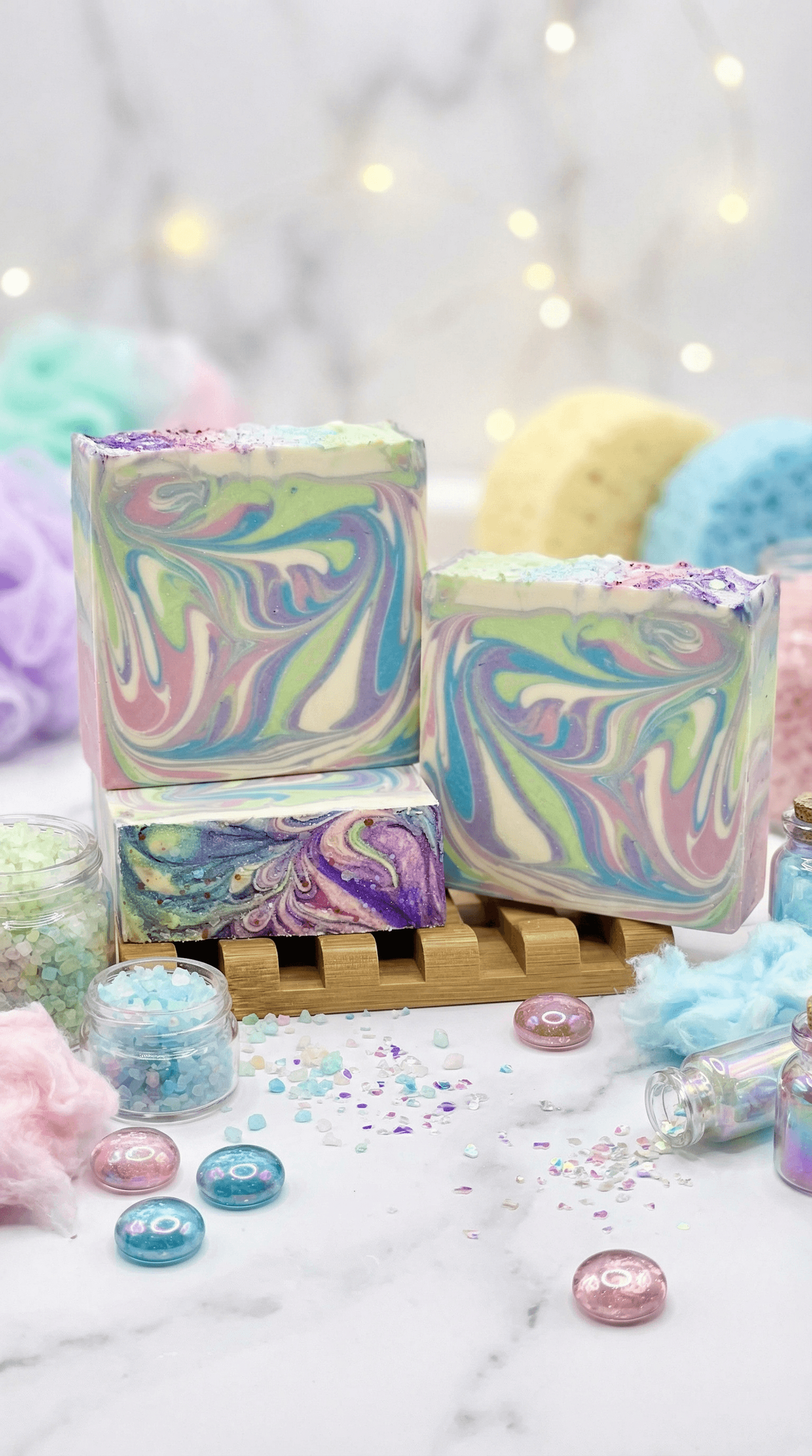 Three handmade soap bars with vibrant pastel swirls in purple, pink, yellow, and turquoise, displayed on a wooden soap dish surrounded by bath salts and decorative gems.