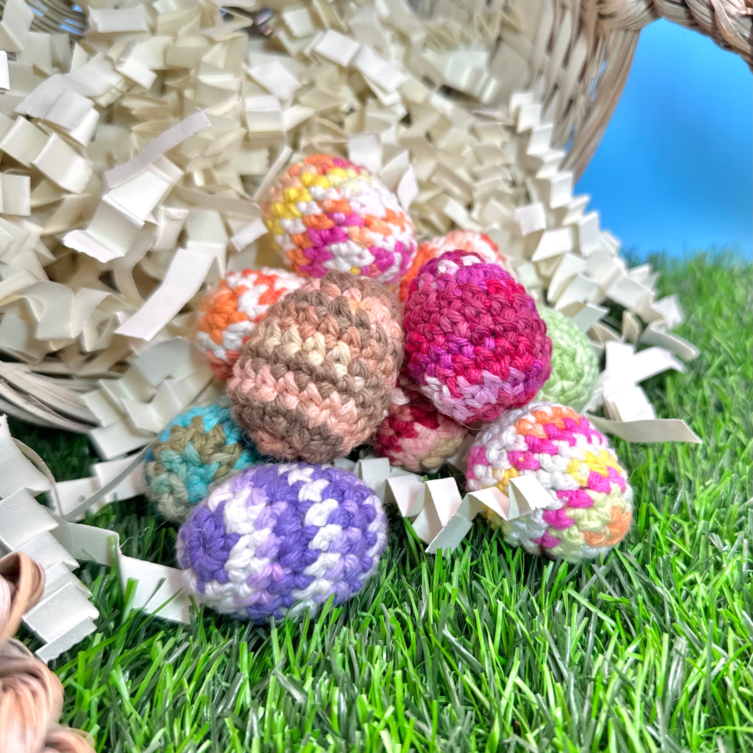 Colorful crocheted eggs in various patterns spilling from a wicker basket onto artificial grass. The handmade eggs feature stripes, solids, and multicolor designs in pink, purple, brown, and rainbow hues.