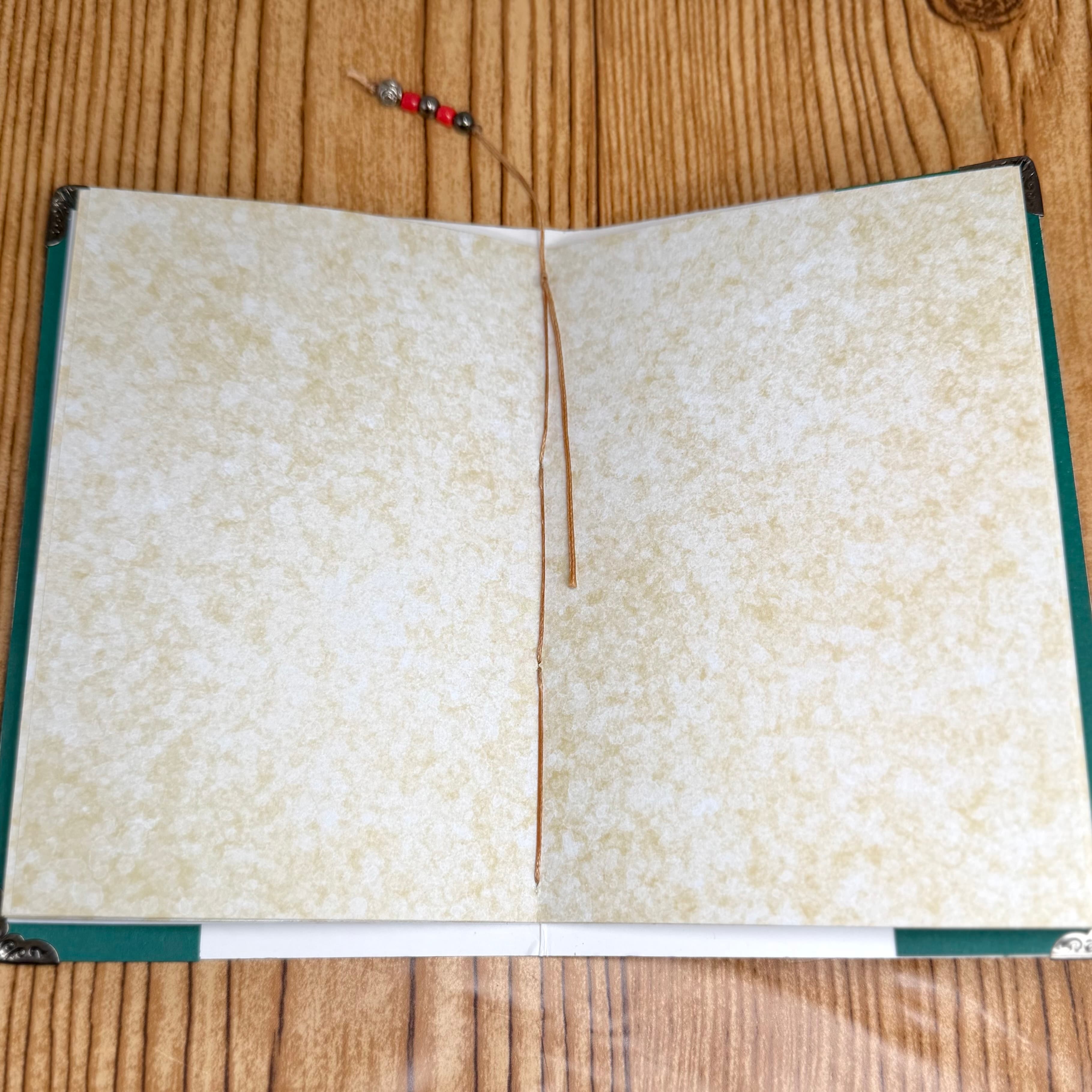 Open handmade journal with cream-colored mottled pages, green borders, and visible hand-stitching down the center. Decorative beaded bookmark rests at top on wooden surface.