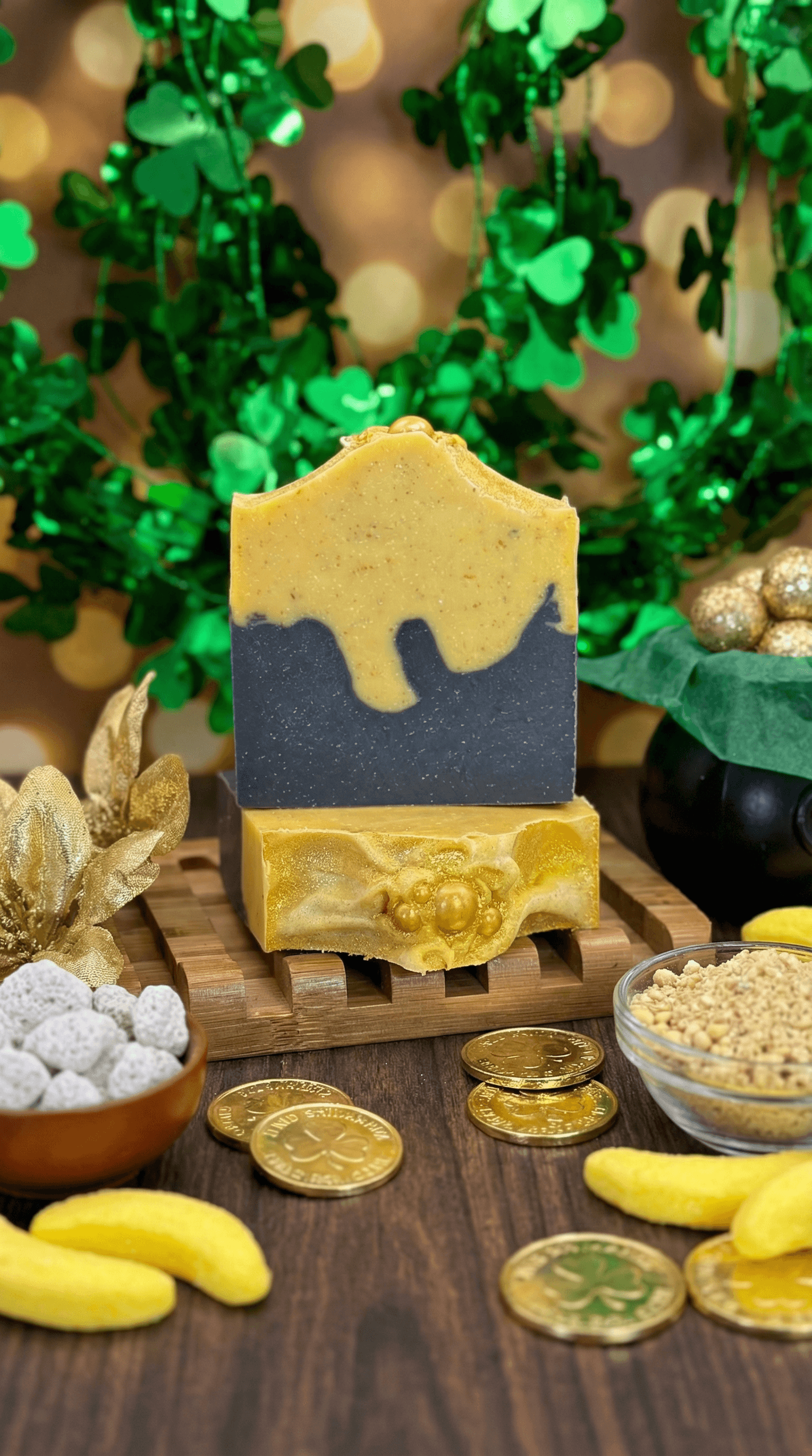 Two-toned bar with shimmering gold top layer dripping over charcoal gray base, displayed on wooden soap dish surrounded by gold coins, shamrock decorations, and yellow marshmallows.