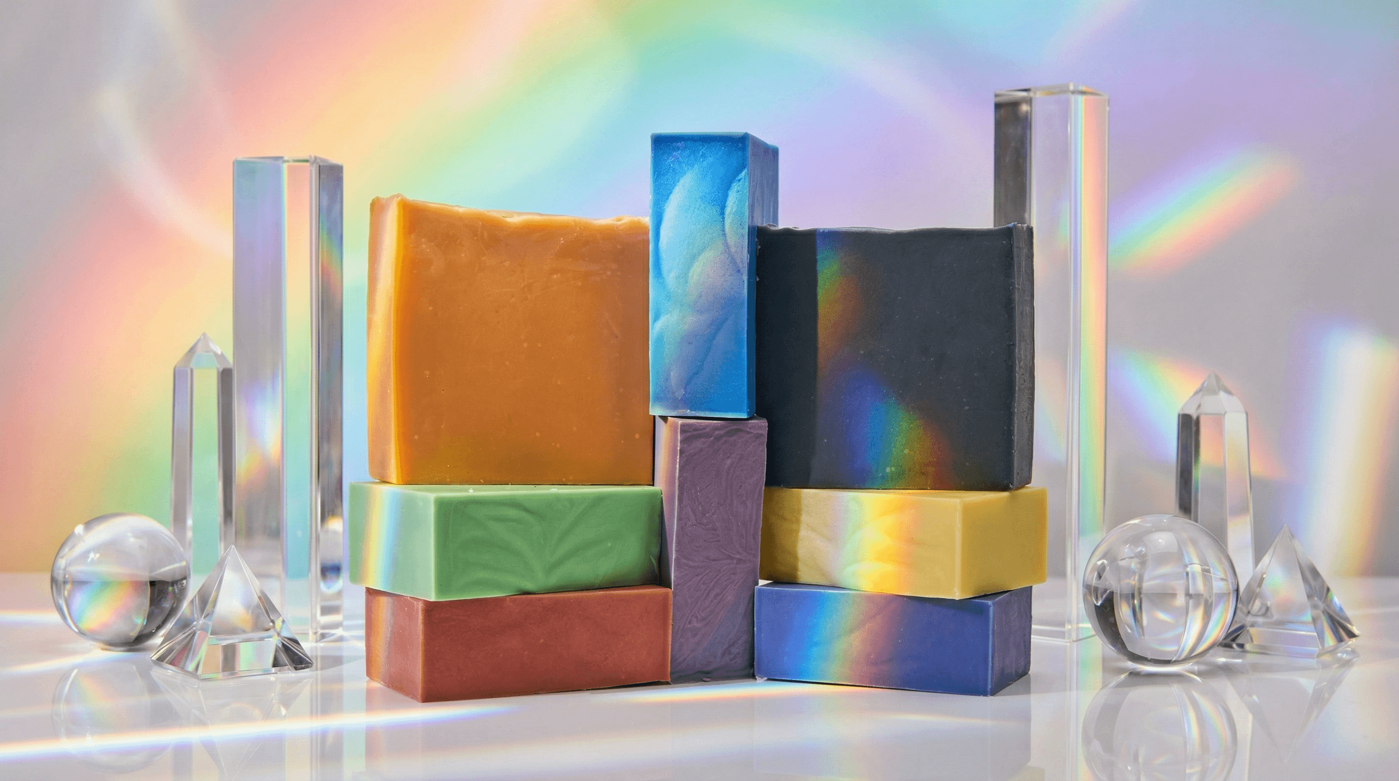 Stacked rainbow-colored soap bars in red, orange, yellow, green, blue, indigo, violet, and black arranged with clear prismatic crystals and spheres creating light refractions.