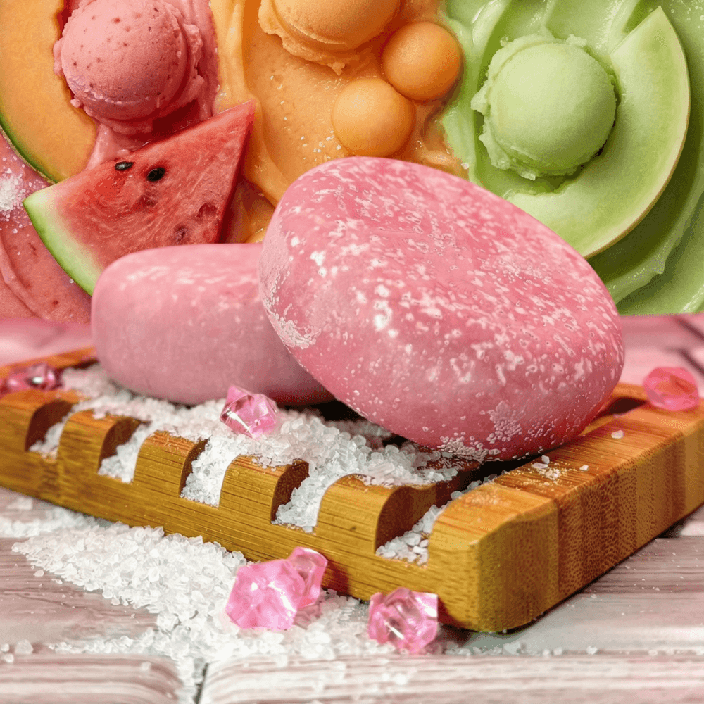 Two large oval pink mochi-textured bath products dusted with white sea salt on bamboo board, surrounded by fresh melon slices, scoops of sorbet, and scattered pink crystals.