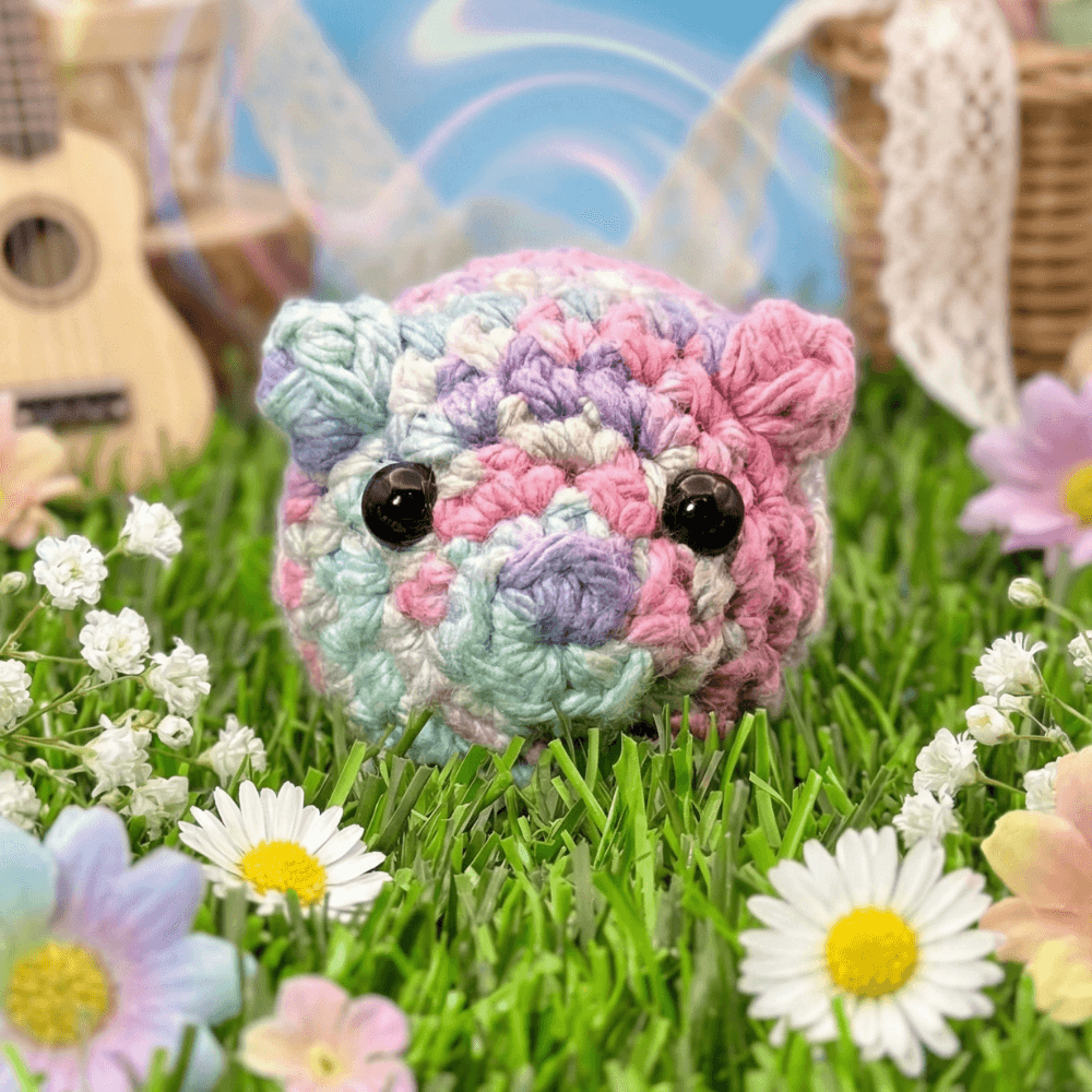Pastel variegated crochet pig sachet in pink, purple, mint and white with black safety eyes, displayed on artificial grass with white daisies and spring decorations.