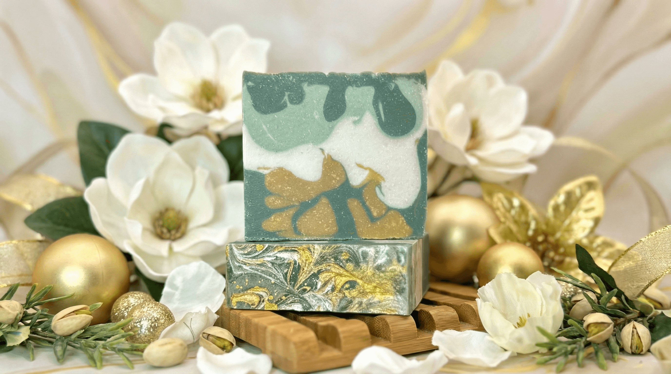 Two stacked handcrafted soap bars with teal green and white swirled design, gold mica accents, and floral pattern on top. Displayed on wooden soap dish surrounded by white magnolia flowers, gold ornaments, and pistachios.