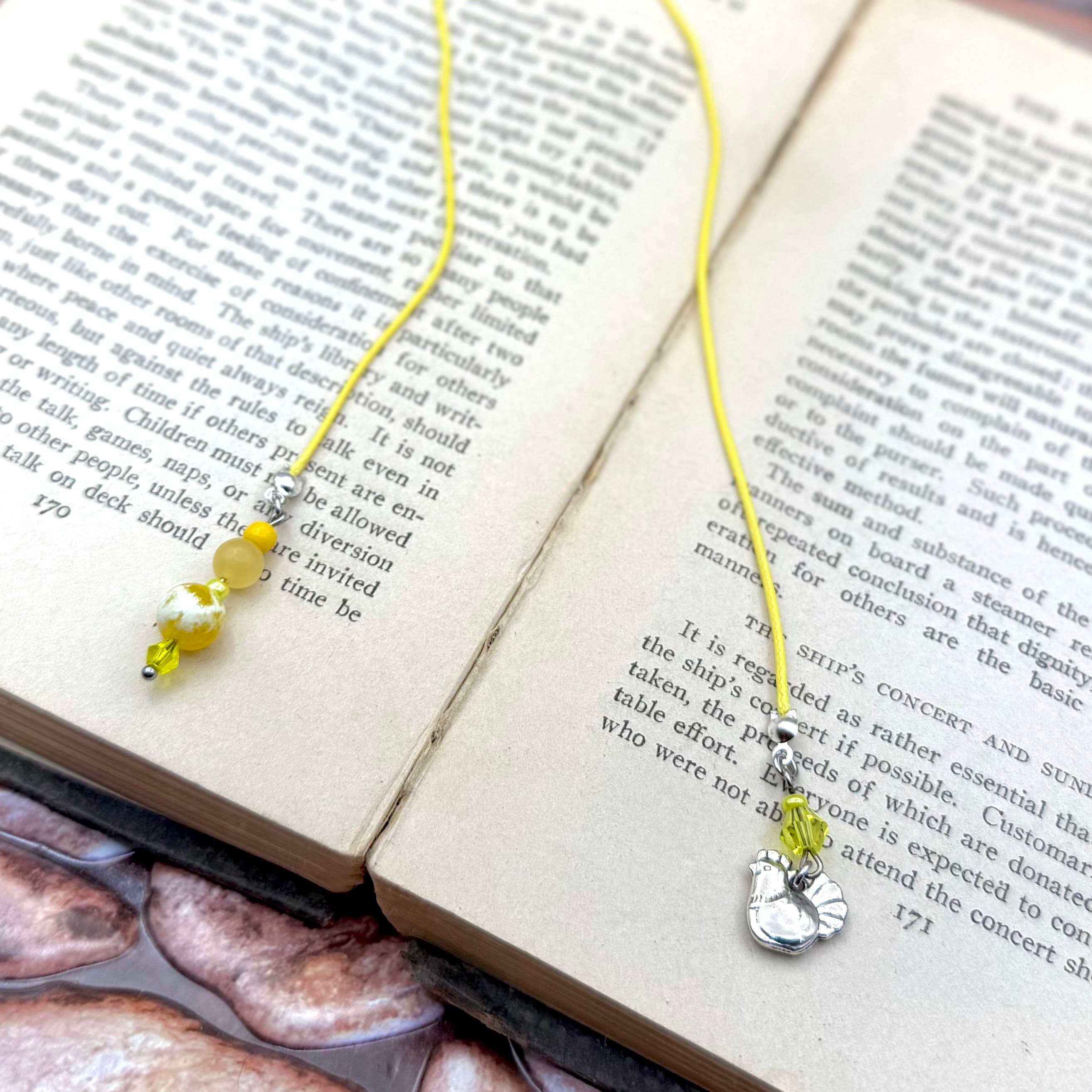 Yellow corded bookmark with hen charm and yellow-white beads resting on open book pages. Two bookmarks shown displaying the design from different angles on vintage text.