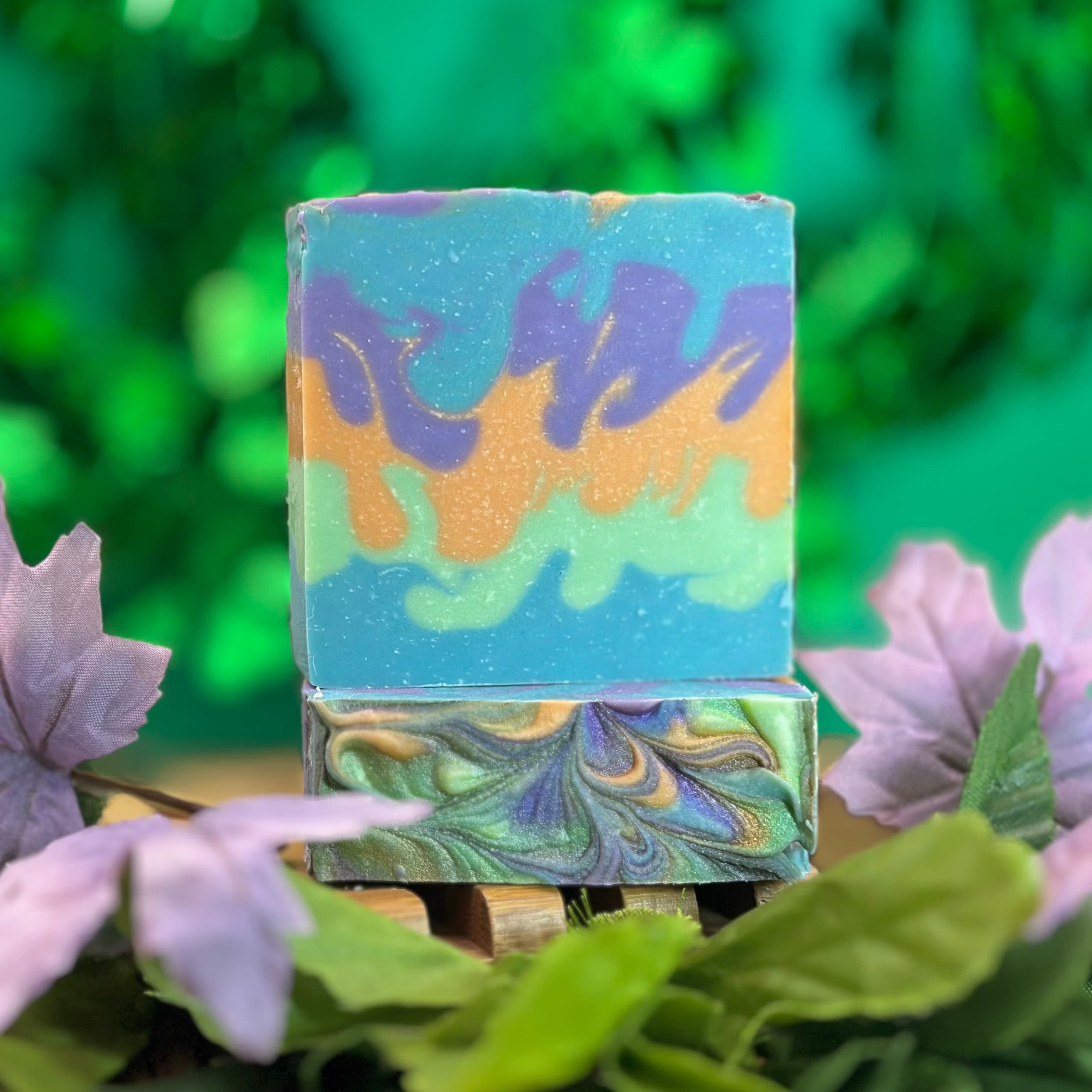 Two stacked handmade soap bars with swirled patterns in blue, purple, orange, and green tones with shimmer, displayed on a wooden soap dish surrounded by purple flowers and green foliage.