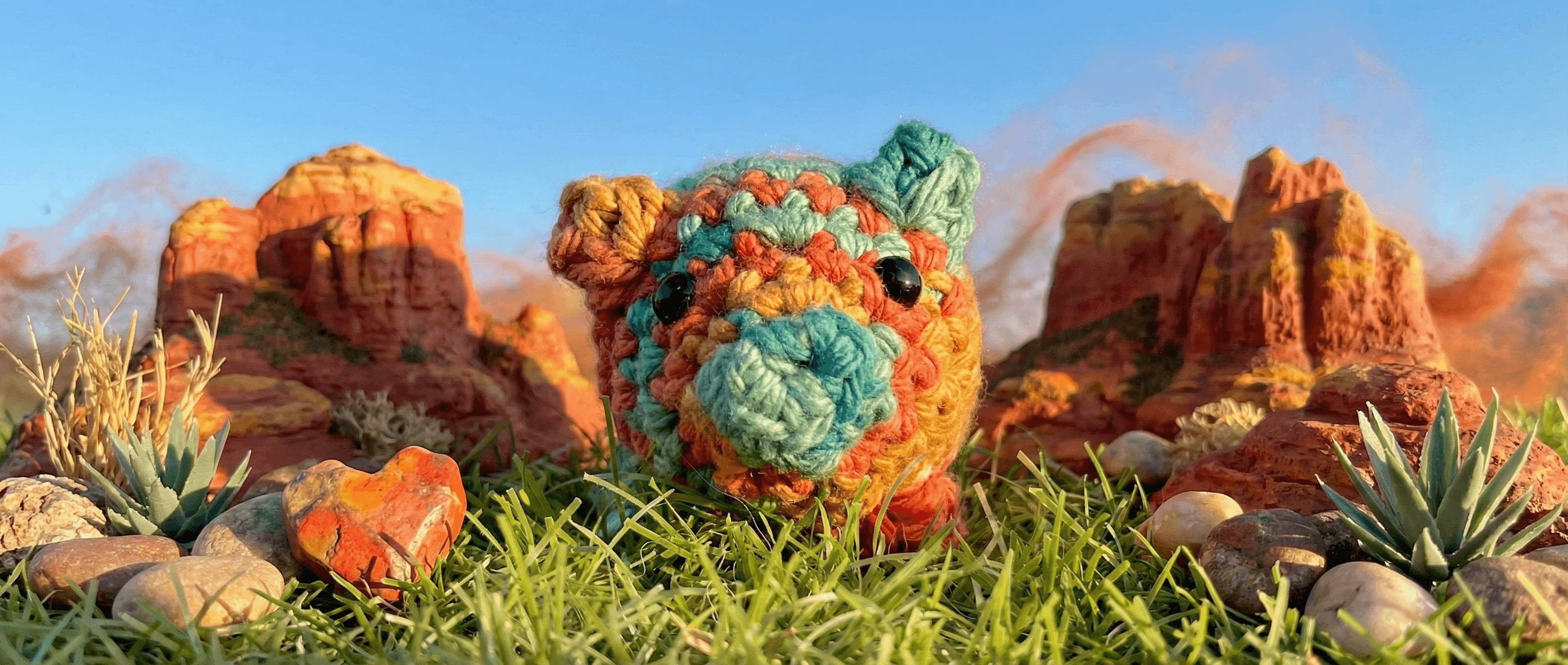 Hand-crocheted pig sachet in multicolored granny square pattern with turquoise snout and ears, photographed on grass with desert red rock formations in background.