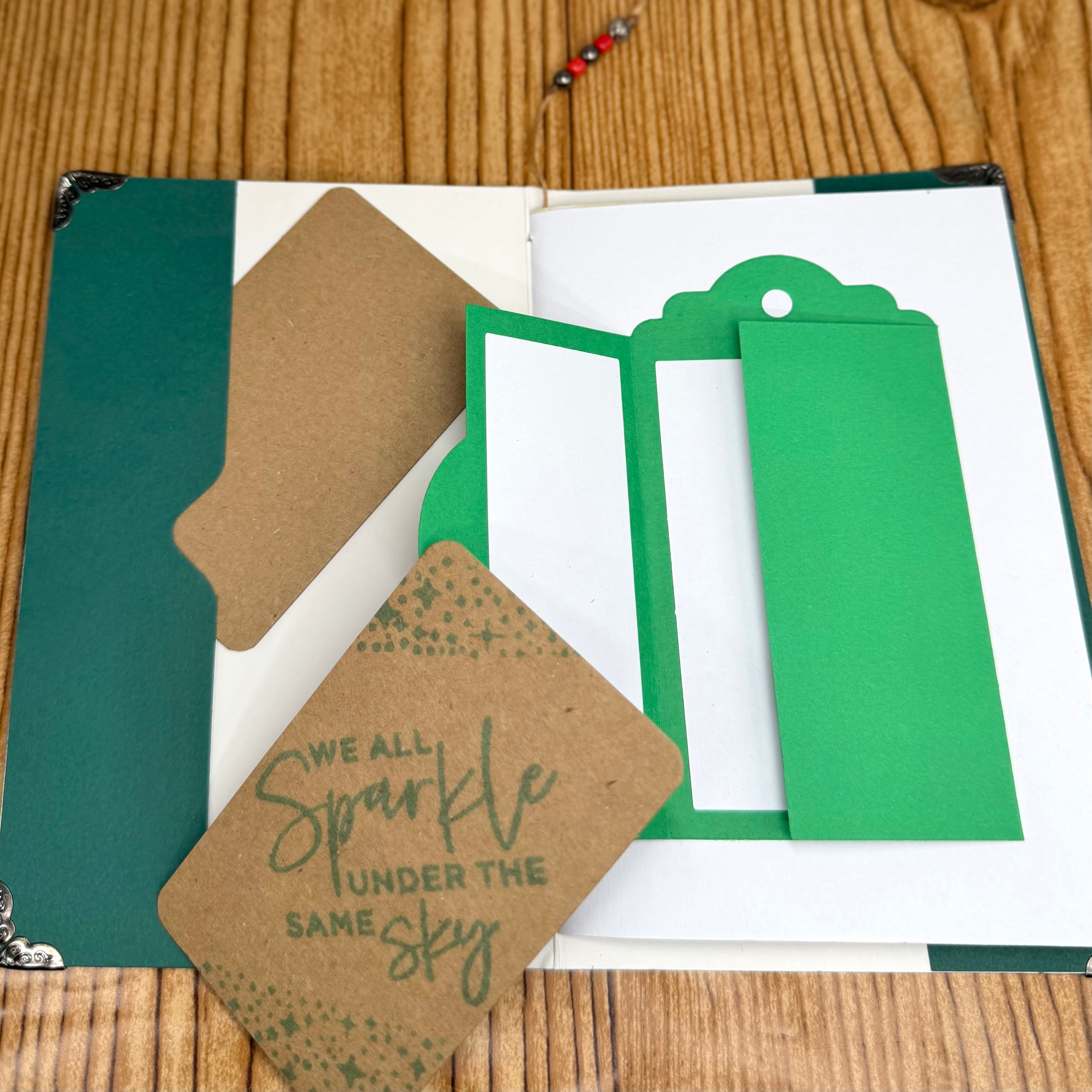 Green paper bookmark and kraft brown affirmation tags with hand-lettered quote on white journal pages, displayed on wood grain surface with decorative beaded accent.