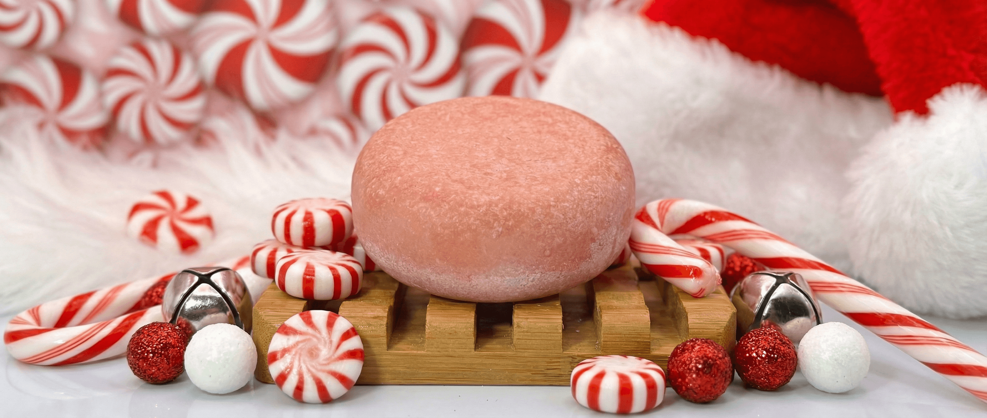 Pale pink oval sea salt scrub bar with sugared texture resting on wooden soap dish, surrounded by red and white peppermint candies, candy canes, and festive holiday decorations.