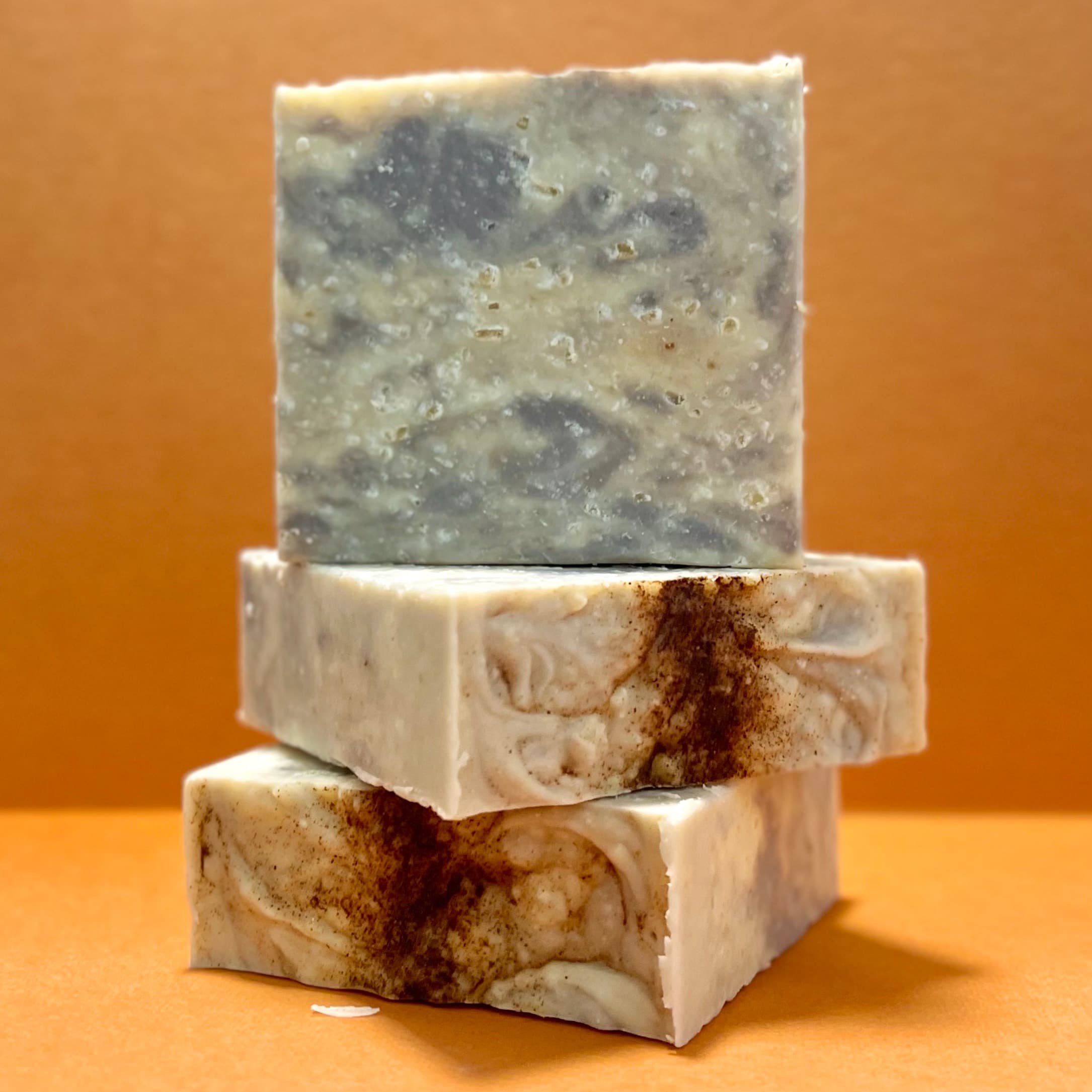 Three stacked bars: top bar is gray-blue marbled, bottom two are cream-colored with brown cinnamon swirls. All bars show textured surfaces with visible exfoliant specks.
