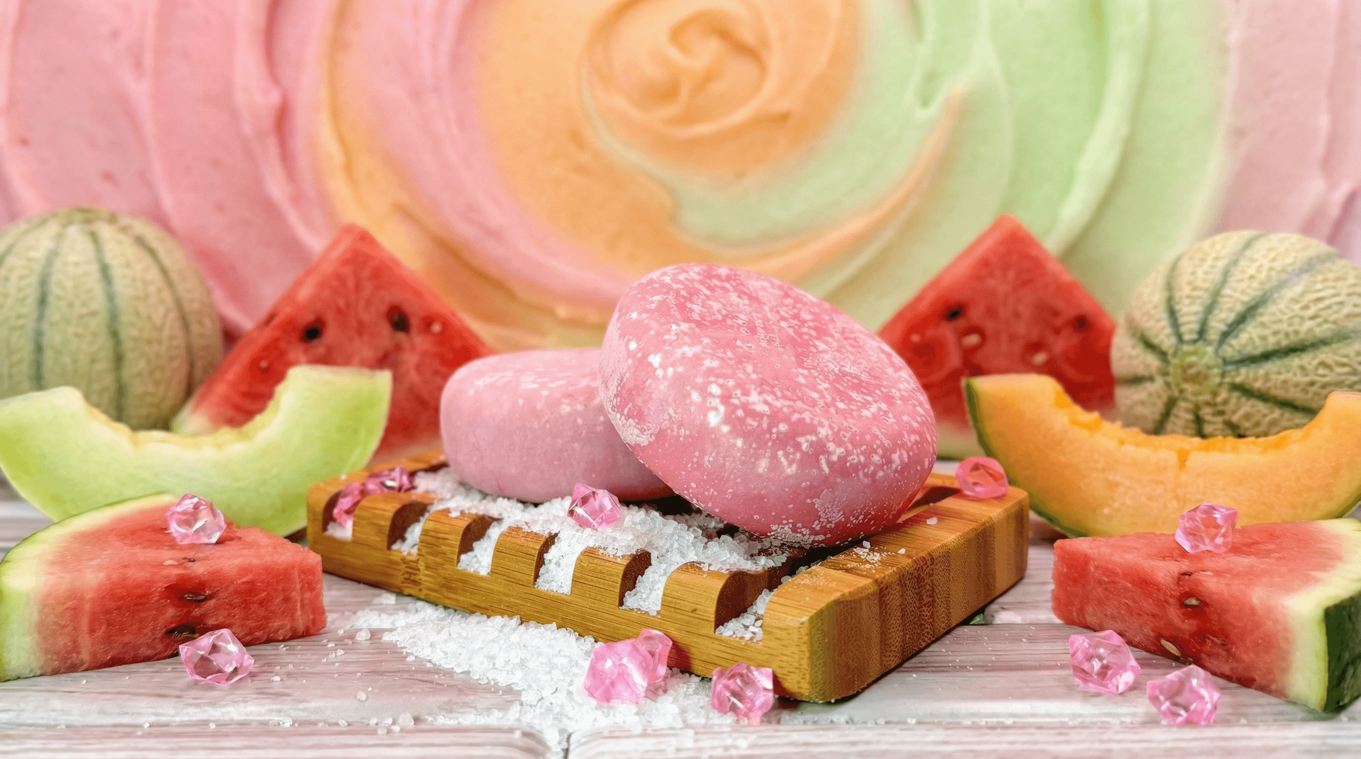 Pink oval sea salt scrub bar with crystallized surface resting on wooden soap dish, surrounded by fresh watermelon and melon slices, pink gem accents, and scattered sea salt.