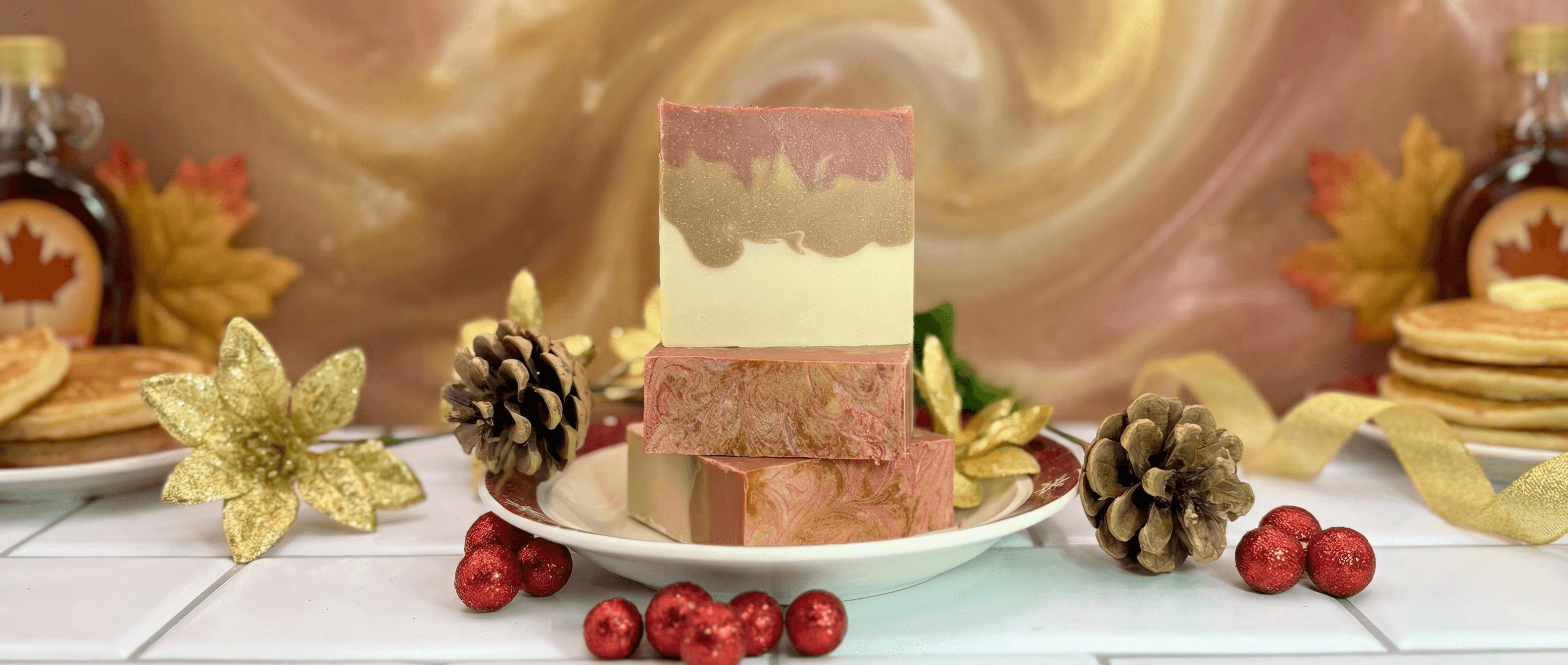 Three handcrafted soap bars in cream, olive green, and mauve pink with swirled marbled texture, displayed on white plate with gold flowers, pinecones, and red ornaments in autumn holiday setting.