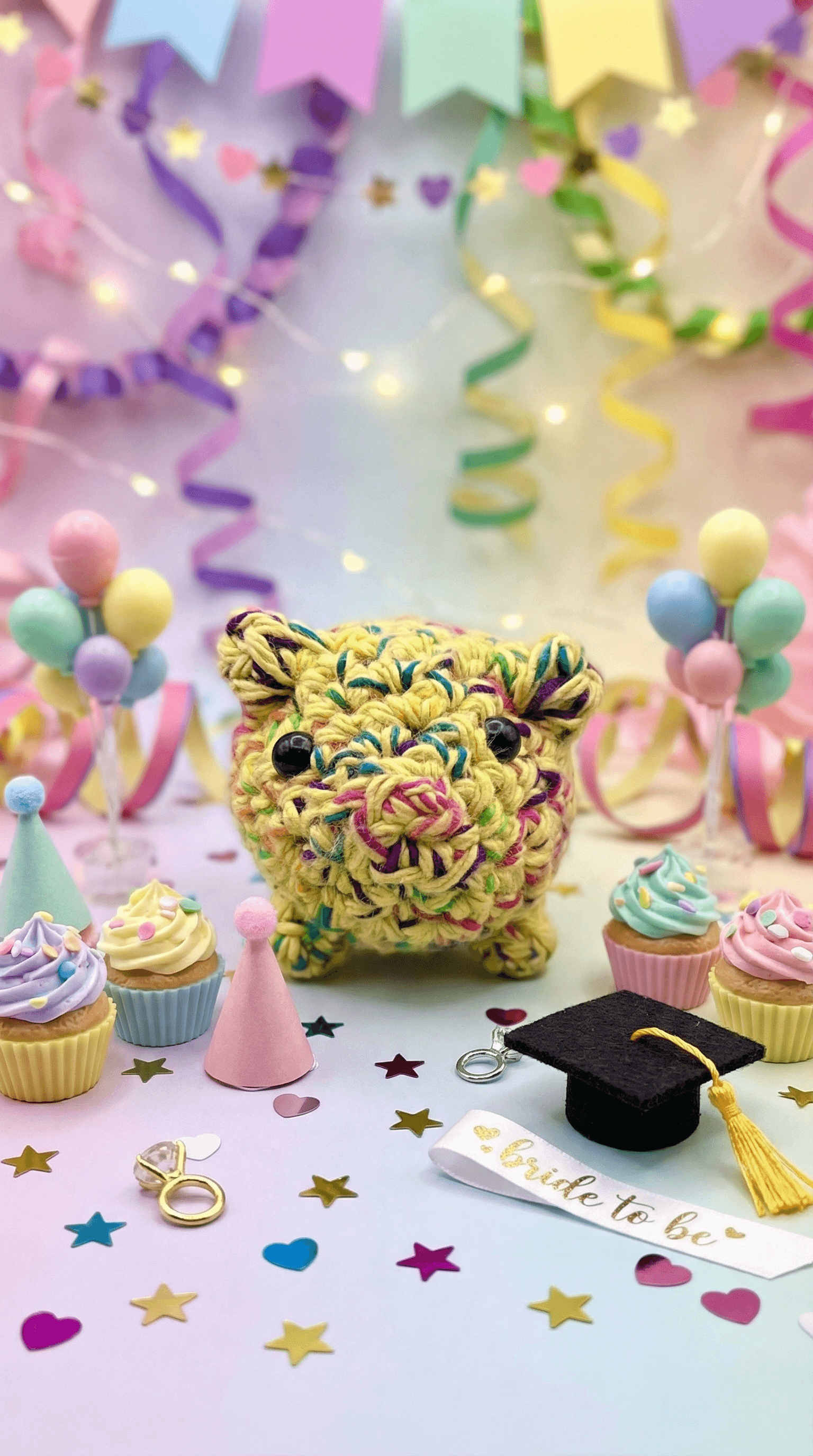 Yellow crocheted pig-shaped sachet with colorful flecks, black bead eyes, and pink stitched snout, displayed among pastel cupcakes, balloons, streamers, and party decorations.