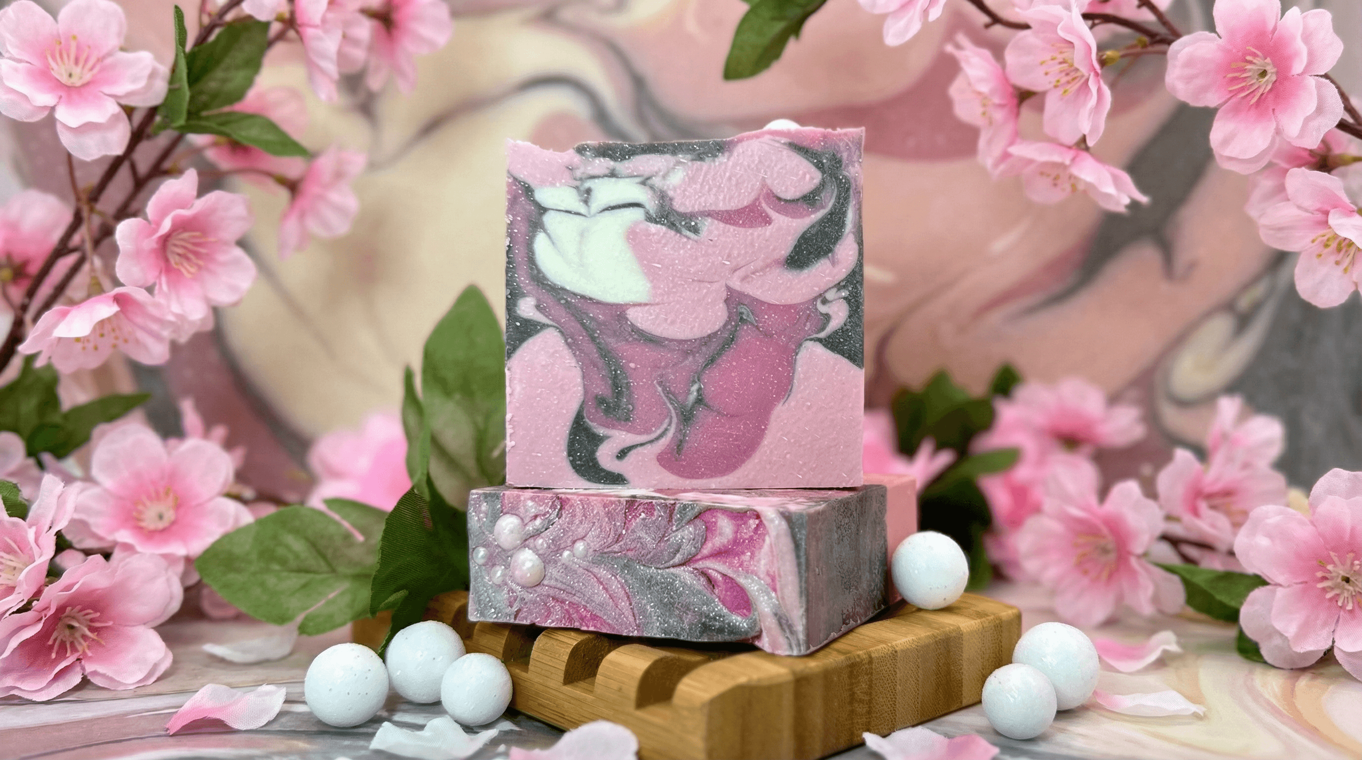 Two stacked bars of marbled pink, mauve, and charcoal gray soap with swirled patterns and light shimmer, displayed on a wooden soap dish with pale blue bath pearls and fresh pink cherry blossoms.