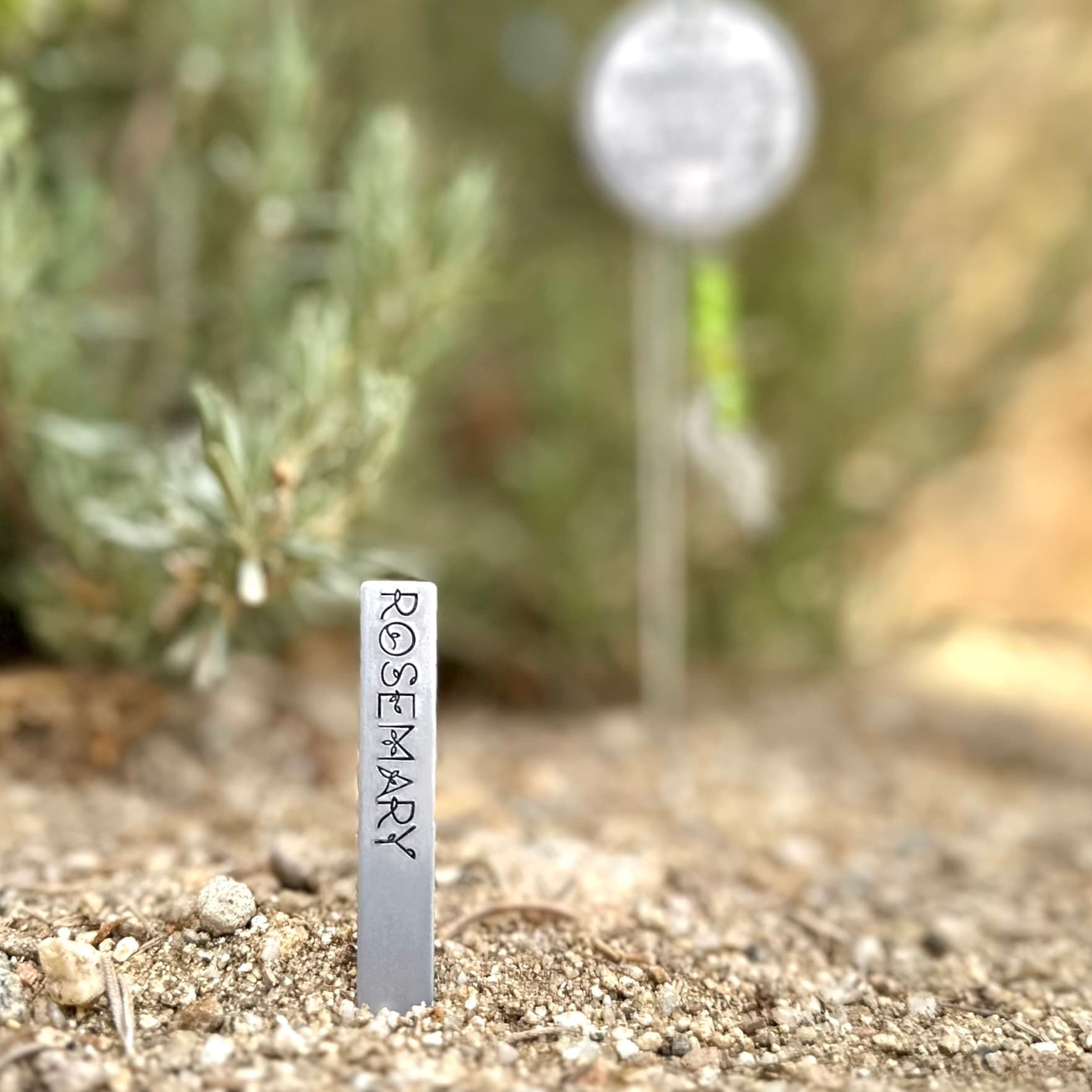 Metal garden stake with 'ROSEMARY' stamped vertically in decorative letters, placed in sandy soil with a small plant and blurred garden marker in background.