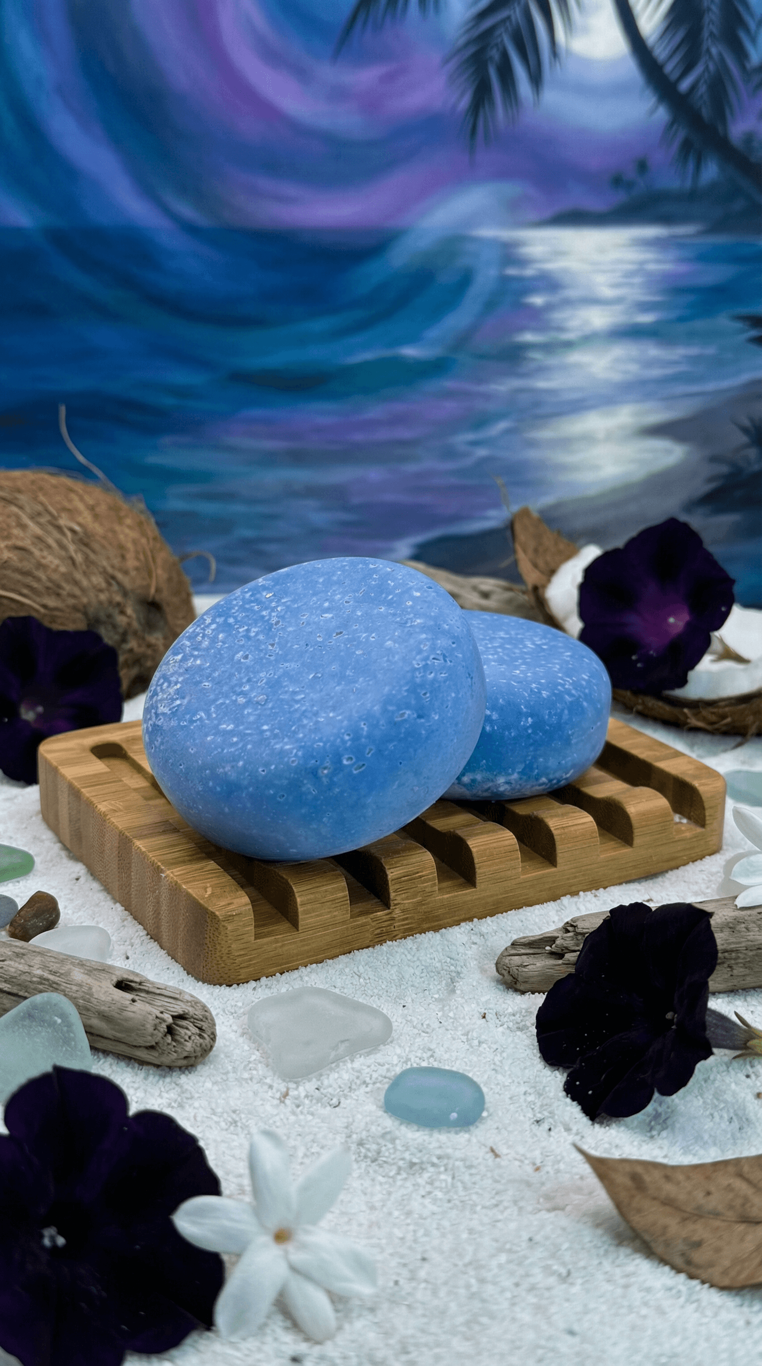 Two smooth blue oval soap bars with speckled texture resting on bamboo soap dish, surrounded by coconut halves, dark purple flowers, and beach glass on white sand.
