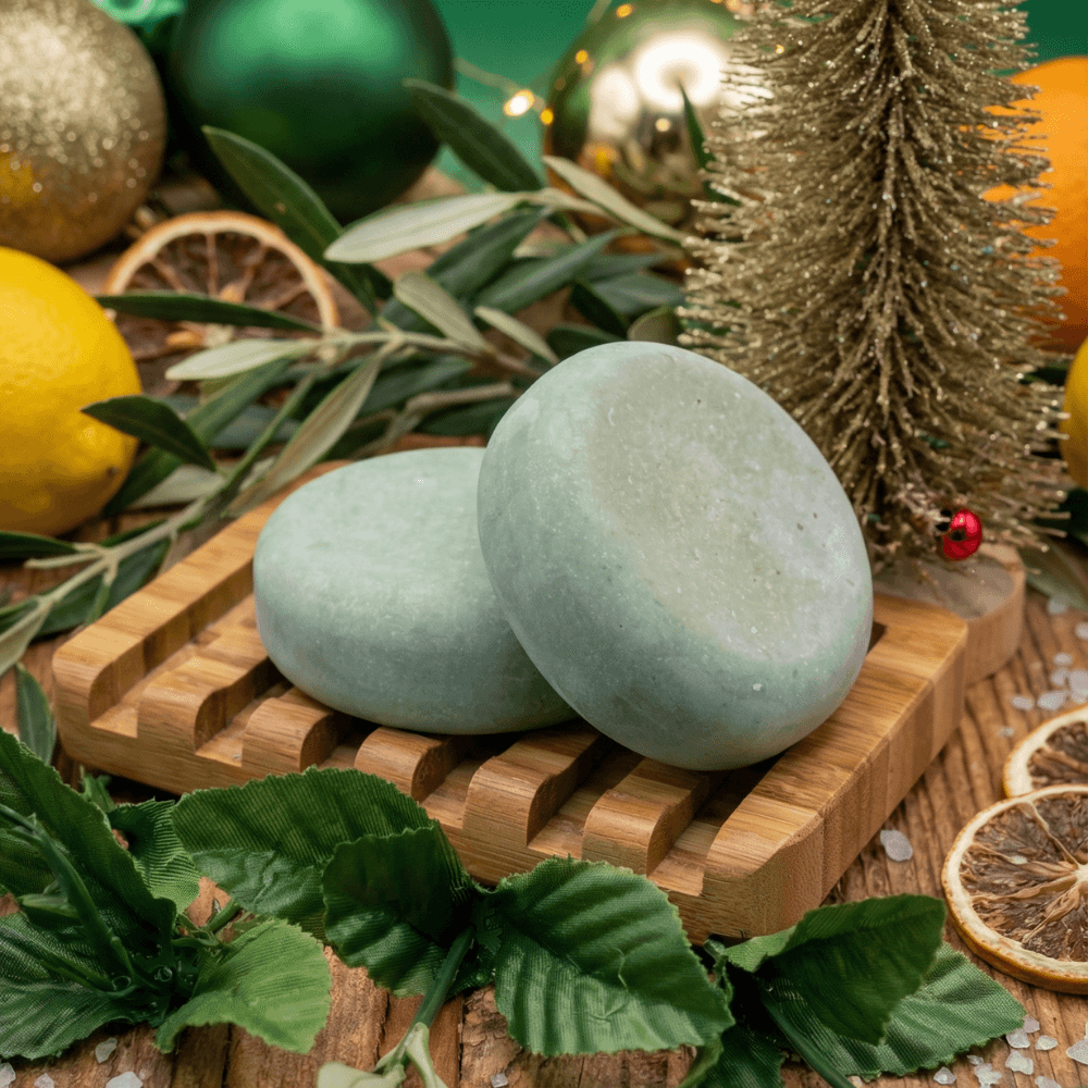 Two smooth, pale sage-green oval soap bars rest on a bamboo soap dish, surrounded by fresh herbs, dried citrus slices, miniature evergreen tree, and holiday ornaments.